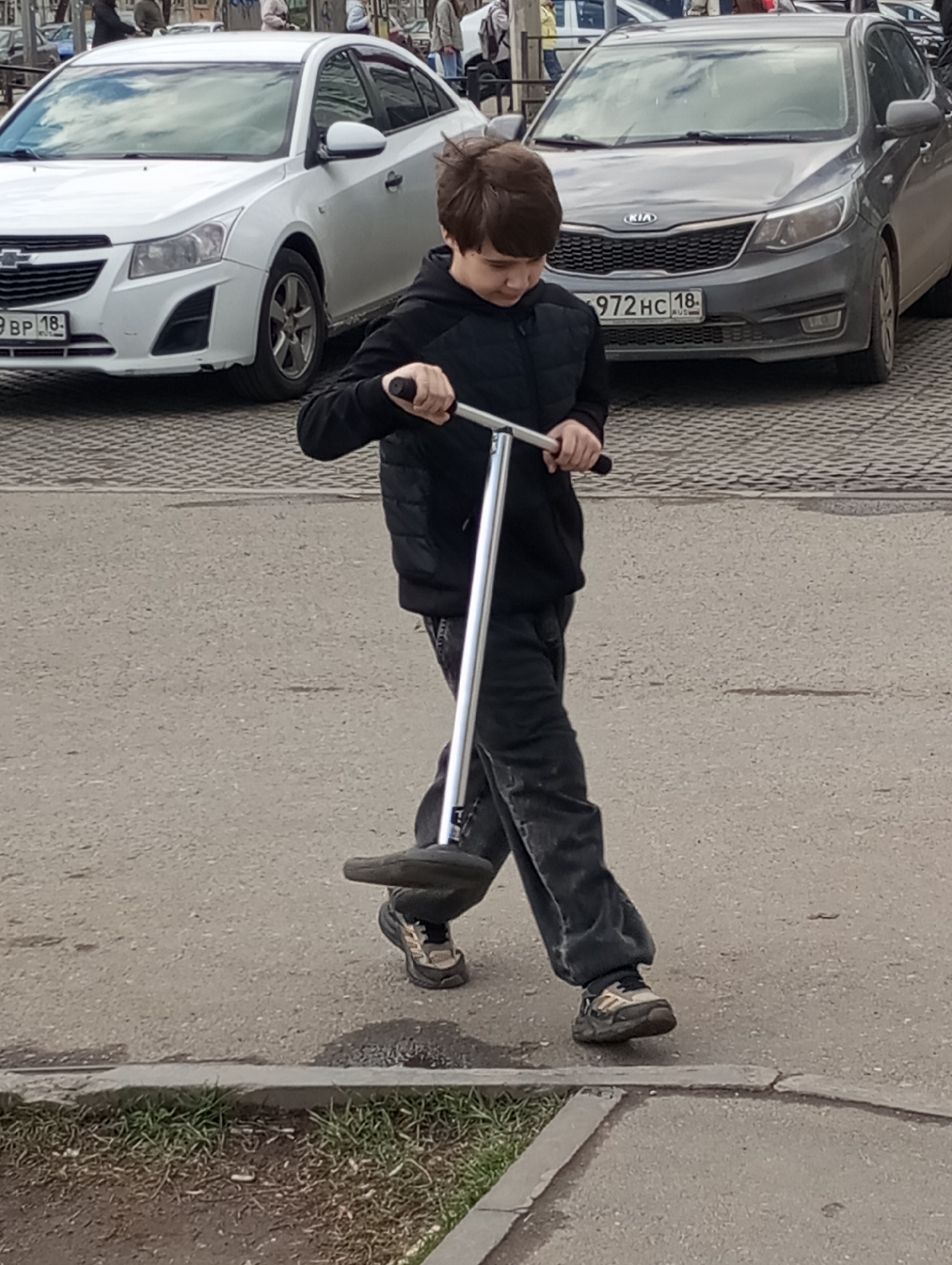 The image captures a young boy with a pogo stick on an overcast day in an urban setting, identified as Izhevsk, Russia. The boy, who appears to be pre-teen, has brown hair and is dressed in a black puffer vest over a dark long-sleeved top, dark jeans, and athletic shoes. He is holding the metallic handlebars of the pogo stick with both hands, looking down at its foot platform, and seems to be in motion, perhaps walking with the pogo stick or preparing to use it. His left foot is on the ground, and his right foot is positioned near the pogo stick's base.
The scene is an outdoor area, likely a street or parking lot beside a sidewalk. The ground is a combination of asphalt and concrete. A curb separates the paved area from a narrow strip of grass and dirt, where some debris, including cigarette butts, is visible.
In the background, two cars are parked. On the left is a white Chevrolet Cruze, featuring a Russian license plate "9 BP 18". To its right is a gray Kia Rio with the Russian license plate "972 HC 18". Both license plates show the region code "18", which corresponds to the Udmurt Republic, confirming the location context of Izhevsk. Partially obscured figures of other people can be seen in the distant background, indicating a public space. The lighting is soft and natural, consistent with a cloudy or partly cloudy daytime sky, which is also reflected in the car windows. The overall impression is a candid moment of daily life in a Russian city.