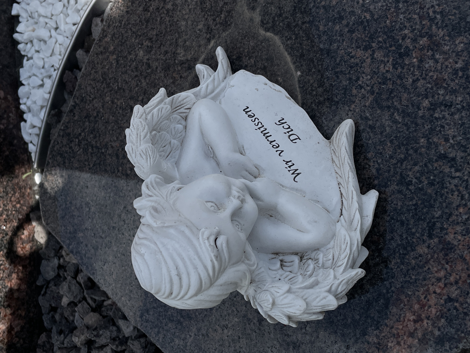 An angel statue is lying on its side on a granite surface. The angel is white and appears to be made of plaster or ceramic. It has its head resting on its folded arms, with wings spread out behind it. On a flat part of the statue, the German phrase "Wir vermissen Dich" is inscribed in black lettering, which translates to "We miss you." The granite surface is dark with flecks of brown and pink. In the background, there are dark gray rocks and some white pebbles. The lighting suggests it is daytime, with shadows cast by the angel.
