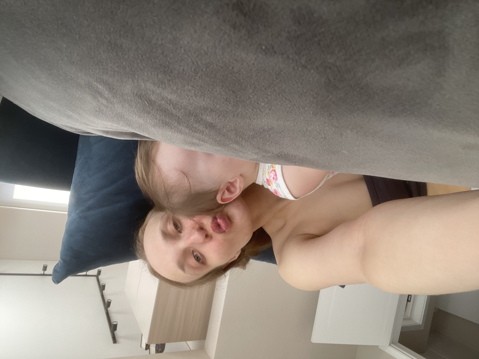 This image captures a candid, intimate moment between a woman and a baby or toddler, set indoors, likely in a residence in Подшивалово, Russia. The photo is taken from a close perspective, with a large grey sofa or cushion partially obscuring the upper portion of the frame.
The primary subjects are a woman, likely the mother, with light hair and fair skin, and a young child. The woman is positioned on her side, looking towards the camera with a playful expression, her lips puckered as if blowing a kiss or making a silly face. She is wearing a dark top, with her bare shoulder visible. Nestled closely to her, with the back of their head and an ear visible, is the baby or toddler. The child also has light hair and fair skin and is wearing a white top with a delicate floral pattern. The child appears to be resting against the woman, possibly on a dark blue pillow that is visible behind the woman's head.
The background features a light-colored wall adorned with a modern black metal light fixture. Further back, a piece of light-colored wooden furniture, possibly a cabinet, is partially visible. The scene is brightly and evenly lit, suggesting it is daytime. The overall emotion conveyed is one of warmth, affection, and playfulness. There is no visible text in the image.