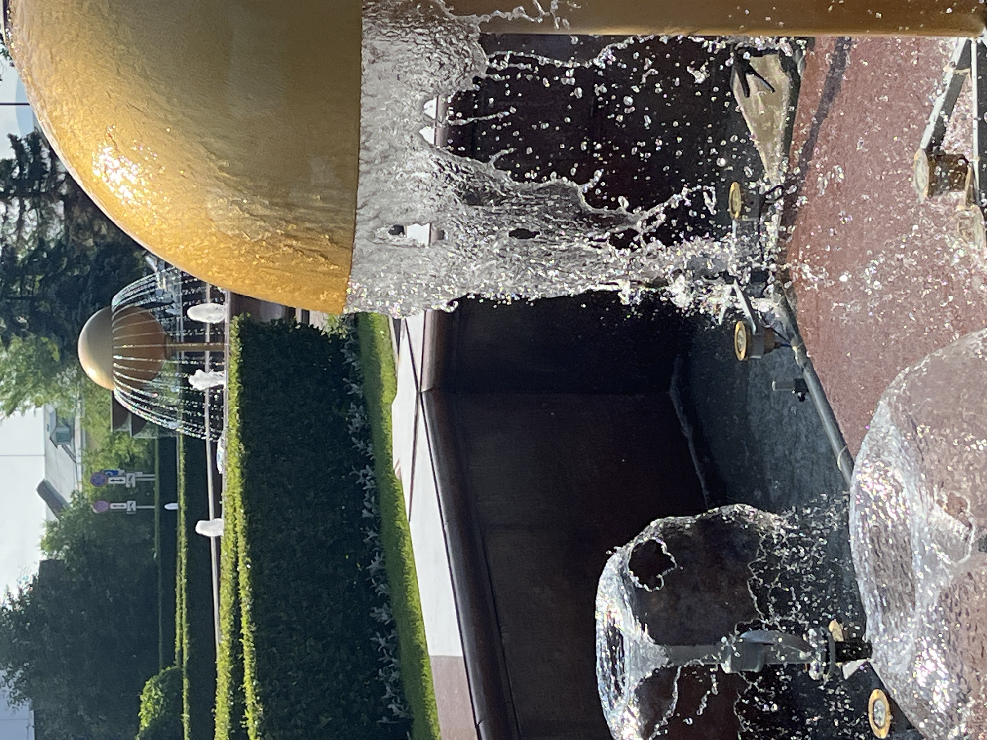 The image shows a close-up of a public fountain in Krasnoyarsk, Russia. In the foreground, water streams from a large, golden, dome-shaped structure and cascades down, creating a shimmering curtain of droplets. To the left, water also flows over two dark, irregularly shaped objects, likely part of the fountain's design. Several smaller water jets are visible, with brass nozzles. The fountain is set within a landscaped area with manicured hedges and grass. In the background, a second, smaller golden dome fountain is visible, also with water spraying from it. Beyond the fountains, trees and buildings suggest an urban setting. The lighting indicates it is daytime.