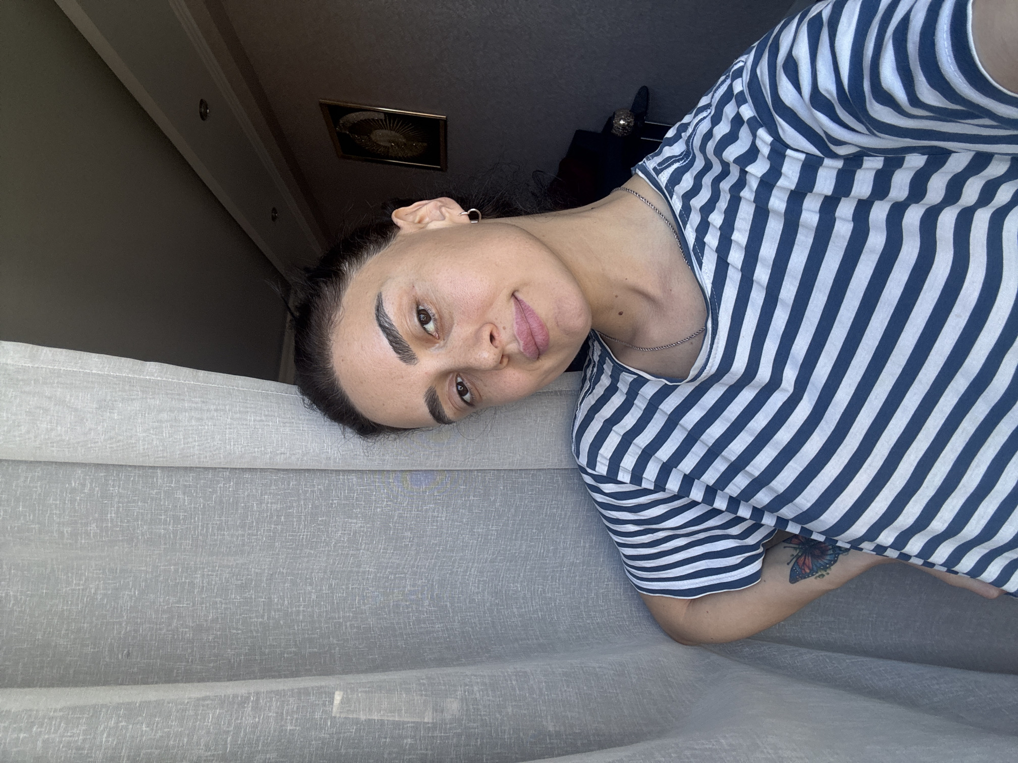 A young woman with dark hair pulled back poses for a selfie indoors, near a sheer white curtain. She is wearing a blue and white striped t-shirt and a silver chain necklace. A small, square framed picture hangs on the wall behind her, which appears to be a dark room with some ceiling lights. The woman has a butterfly tattoo on her left arm. The image was taken in Tbilisi, Georgia.