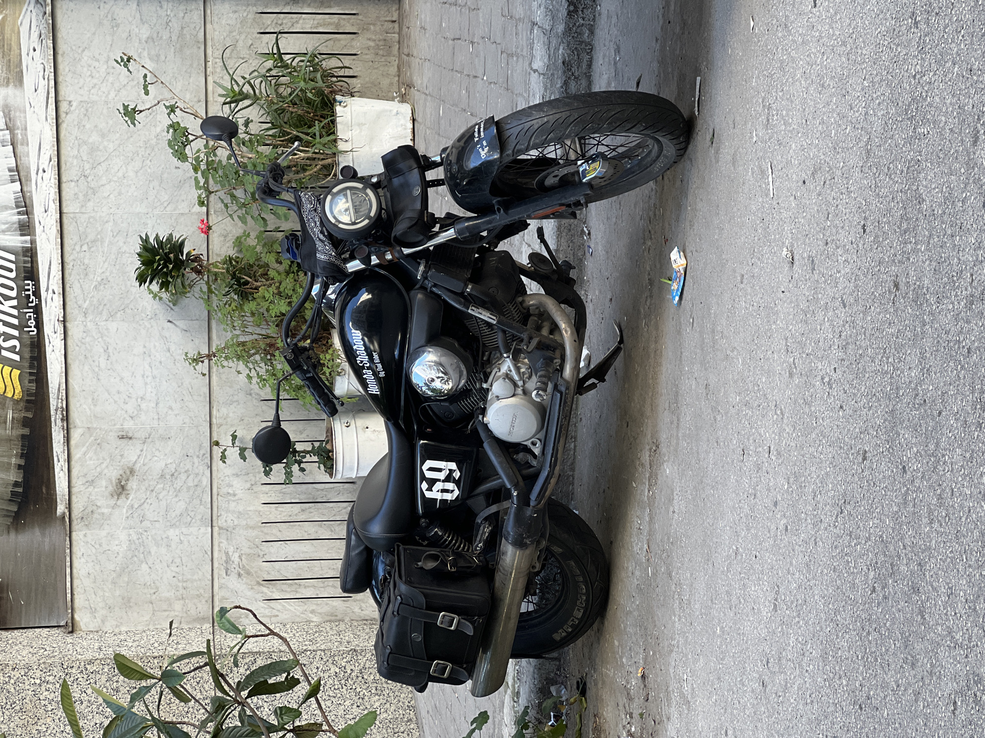 A black Honda motorcycle is parked on the sidewalk in a city environment. The motorcycle has a distinctive number "69" on its side, leather saddlebags, and custom modifications. It is positioned next to a building with marble-like panels and a sign with Arabic script. The background also features various plants and greenery in containers and growing on the sidewalk. The pavement shows typical wear and tear with minor debris. The overall setting suggests an urban environment in Tanger طنجة, Morocco.
