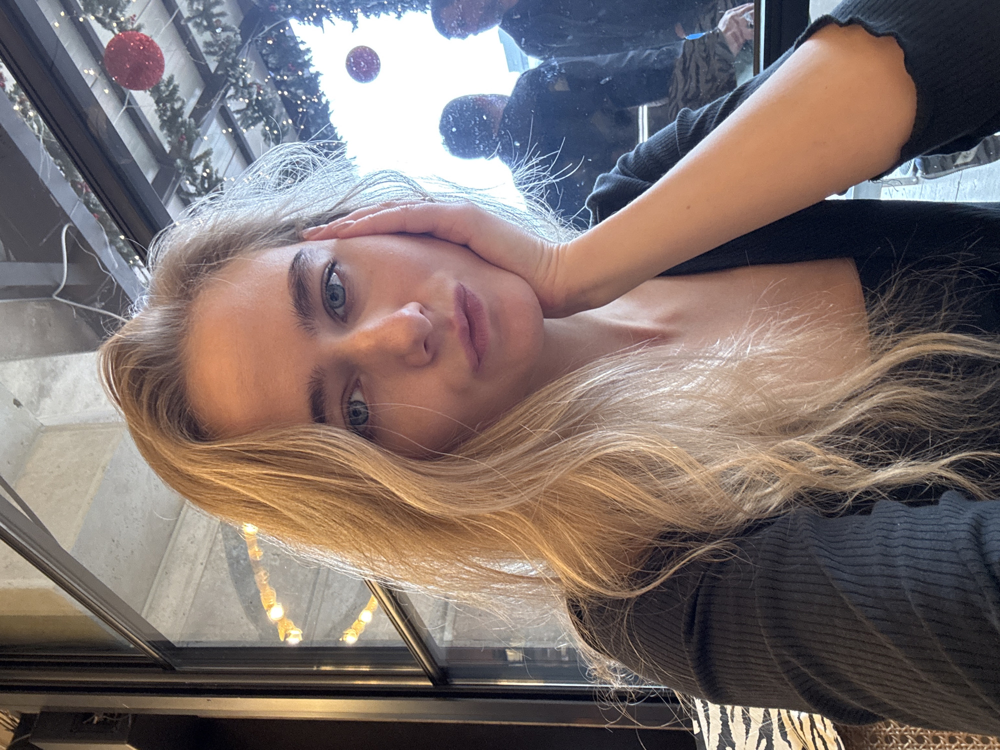 A selfie taken by a fair-skinned young woman with long blonde hair and blue eyes, indoors. The woman has her hand on her cheek and is wearing a black ribbed shirt. A reflection in a window behind her shows holiday decorations, including red ornaments and greenery, and indistinct figures. It is in an unknown location.