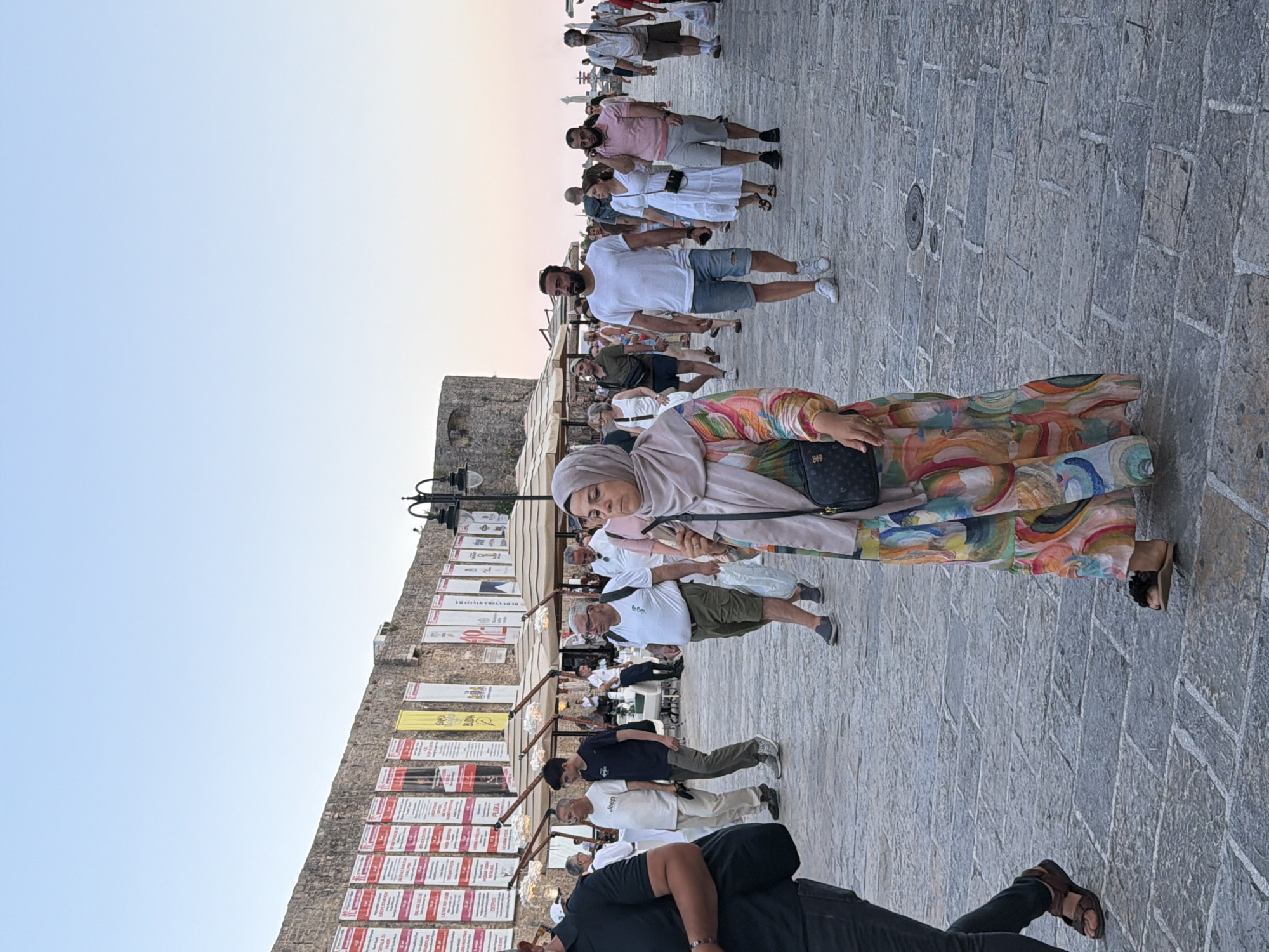 The image depicts a bustling street scene in Budva, Montenegro, likely near the Old Town walls. The paving stones suggest a historic promenade. On the left, a person in a black t-shirt and jeans walks away from the camera. In the center foreground, a woman wearing a brightly colored, patterned dress and a light-colored hijab walks towards the camera, carrying a black crossbody bag. Behind her, a crowd of people of various ages and genders are walking in both directions. Many are dressed in casual summer attire, including shorts, t-shirts, and dresses.
In the background, a stone wall is visible, adorned with numerous banners and posters, possibly advertising events or local businesses. Several outdoor cafe seating areas with umbrellas are set up along the wall. The sky above is a clear, pale blue, transitioning to a hint of orange near the horizon, indicating it might be late afternoon or early evening. Streetlights with intricate designs are mounted on poles, adding to the ambiance of the location. The overall impression is one of a lively tourist area.