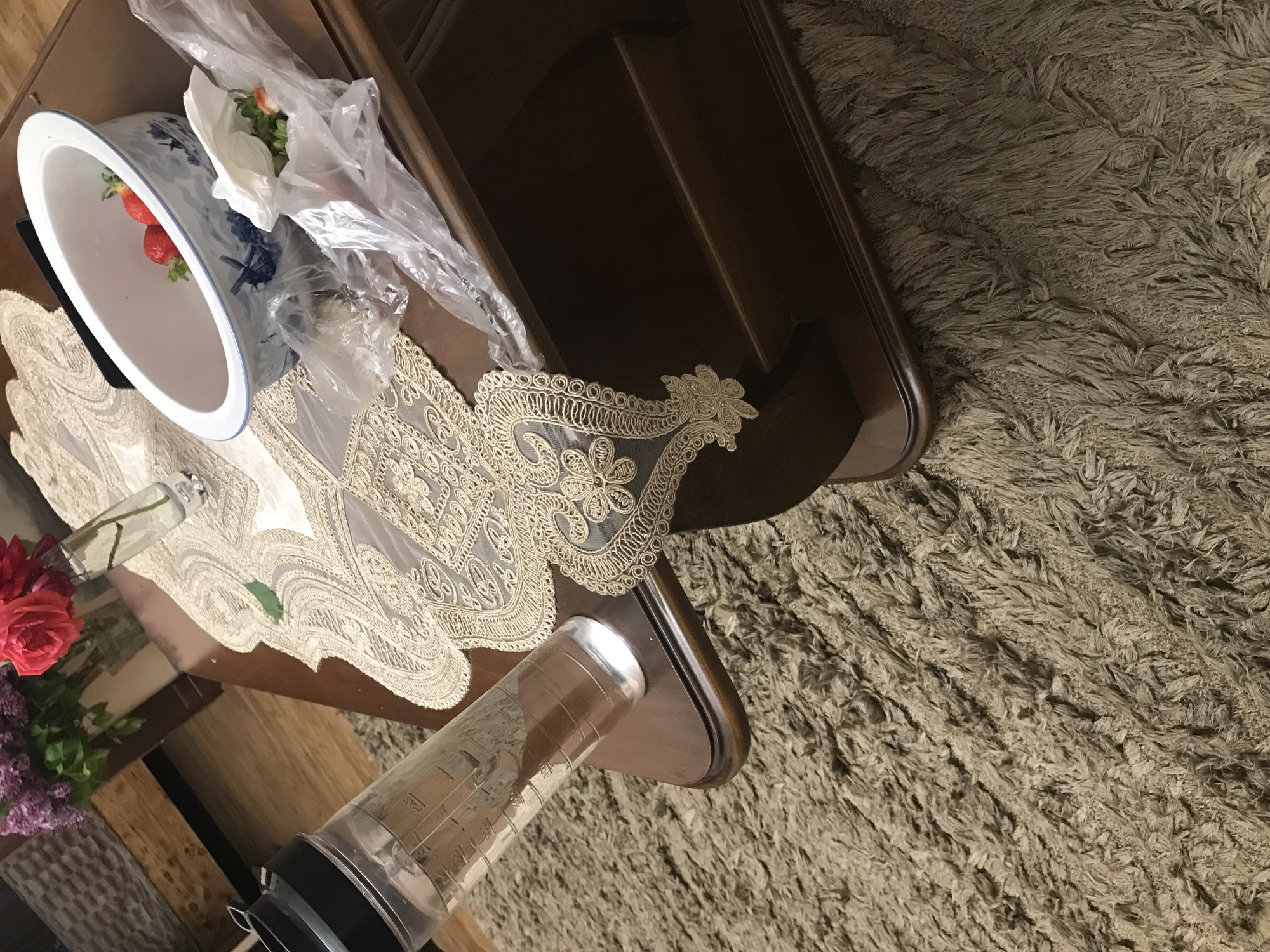 The image shows a high-angle view of a scene on a brown wooden table, partially covered by an ornate, cream-colored lace doily. On the left side of the table is a white bowl with a blue floral design, containing strawberries. Next to the bowl is a plastic bag containing white flowers. A bouquet of roses and lilacs are in a vase. A clear plastic container with black accents is positioned on the edge of the table. The table is placed on a thick shag rug with neutral colors and is located in Yerevan, Armenia.