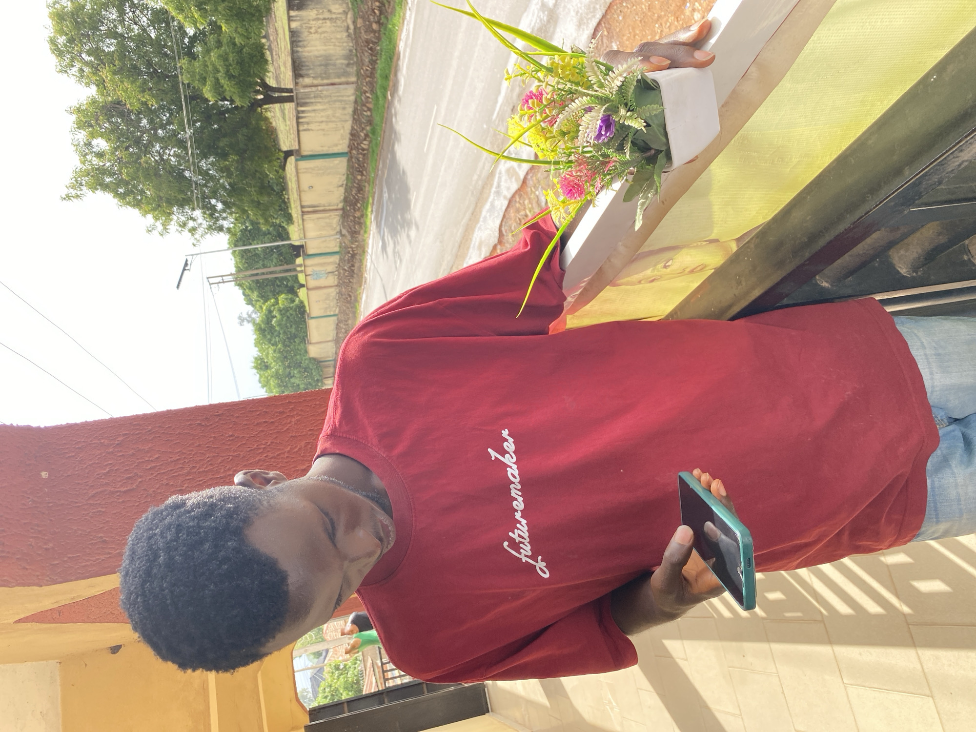 A man stands outside, possibly in Bukuru, Nigeria, holding a teal smartphone. He is wearing a red t-shirt with "futuremaker" written on it and blue jeans. His hair is short and dark. Next to him is a potted arrangement of flowers in a white square pot, resting on a painted railing. The background shows a street scene with a wall and trees.