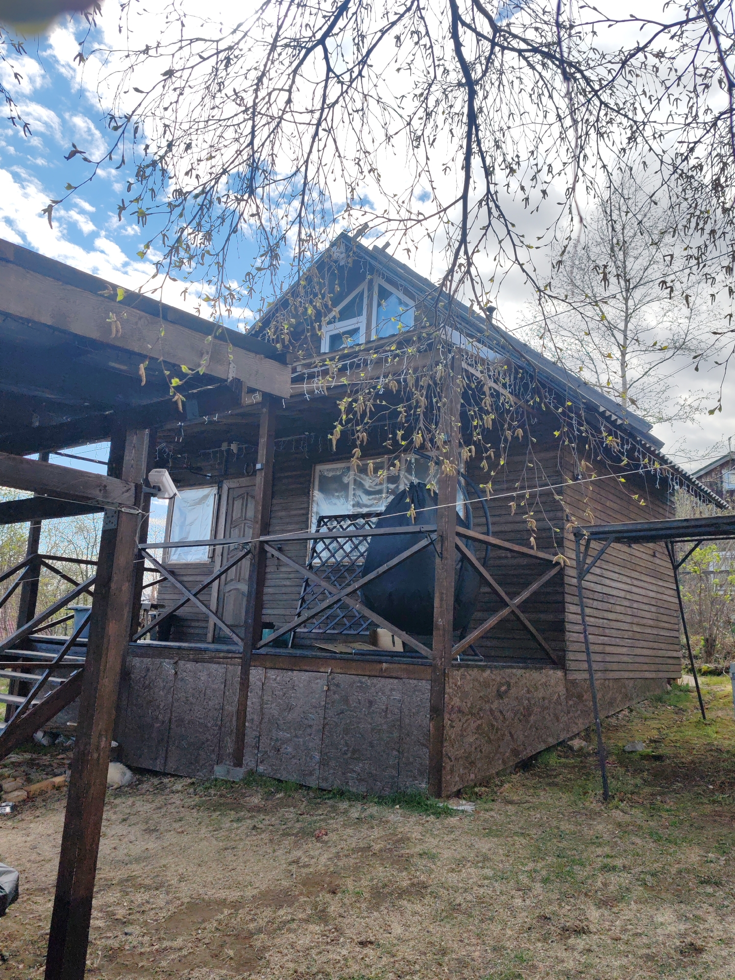 The image shows a wooden house with a porch, likely a dacha or summer house, located in a grassy area with some trees. The house has a dark wood exterior with a gabled roof. A porch with wooden railings extends from the front and side of the house. The siding beneath the porch appears to be OSB. The windows are covered with curtains. The sky is partially cloudy, with visible blue patches. Bare tree branches are visible in the foreground, partially obscuring the view of the house. The location is Koryaki, Russia. There are no people visible in the image.
