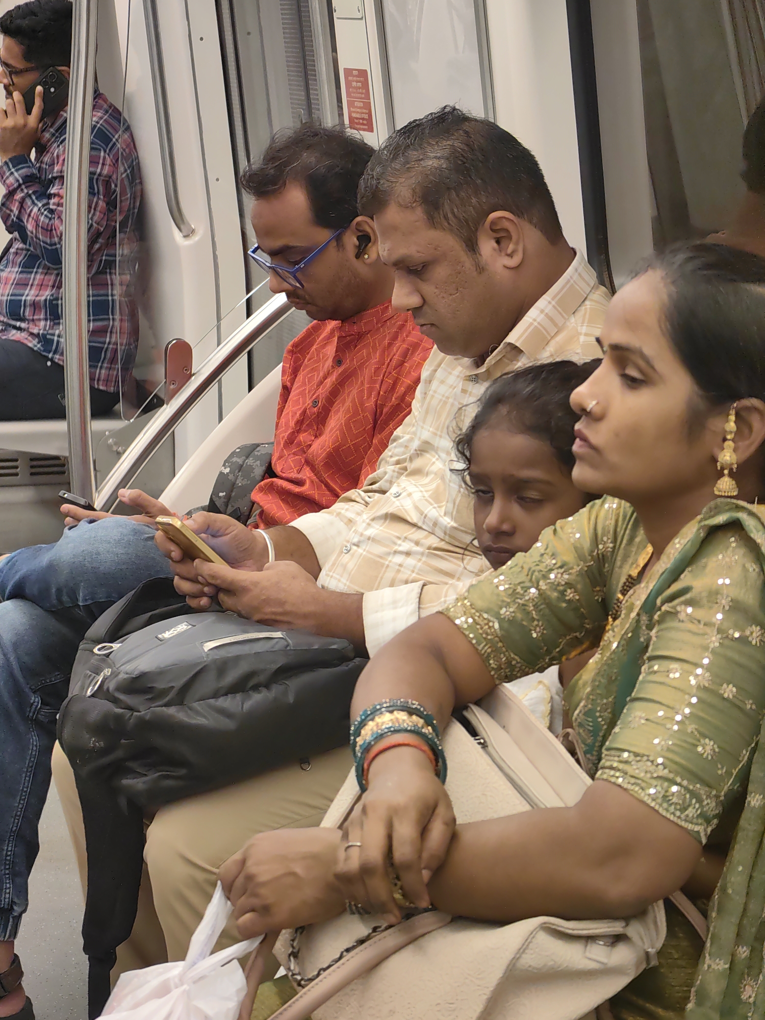 An overhead shot shows several people sitting on a train, possibly the Mumbai Metro, in India. A man with glasses and a plaid shirt is on his phone, while another man in a patterned shirt looks down. A woman in a green outfit with elaborate jewelry sits beside a young girl, and a third woman appears to be holding a white plastic bag. The environment is a train carriage with metal poles and a reflective wall.
