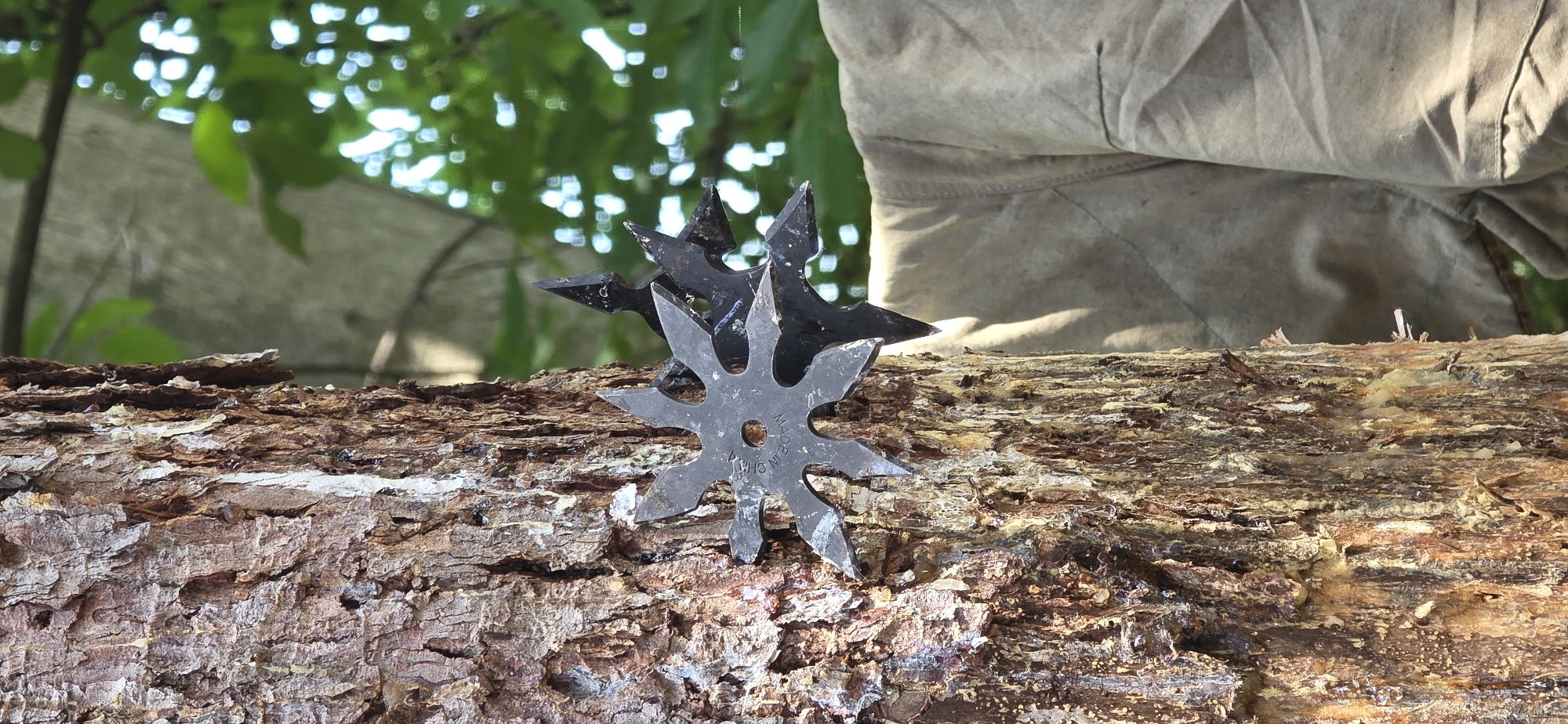 This image, observed in the city of Partizanin, Bulgaria, depicts a close-up, vertical view of a tree trunk with multiple throwing stars (shuriken) embedded within its bark. The most prominent throwing star, situated in the mid-left, is six-pointed, dark gray, and features a central circular perforation. Partial text "MADE IN C" is visible on its surface. Behind and to the right of this star, at least two additional, darker-toned throwing stars are partially visible, also lodged in the bark. The tree trunk exhibits thick, fissured, reddish-brown bark, with substantial amounts of white and yellowish resin or sap adhering to its vertical crevices. The background, to the right of the tree, comprises brightly lit, out-of-focus green foliage displaying distinct circular bokeh. Below the foliage, a segment of light tan textile material, possibly a piece of clothing or canvas, with visible seams and folds, is present. No individuals are depicted in the scene.