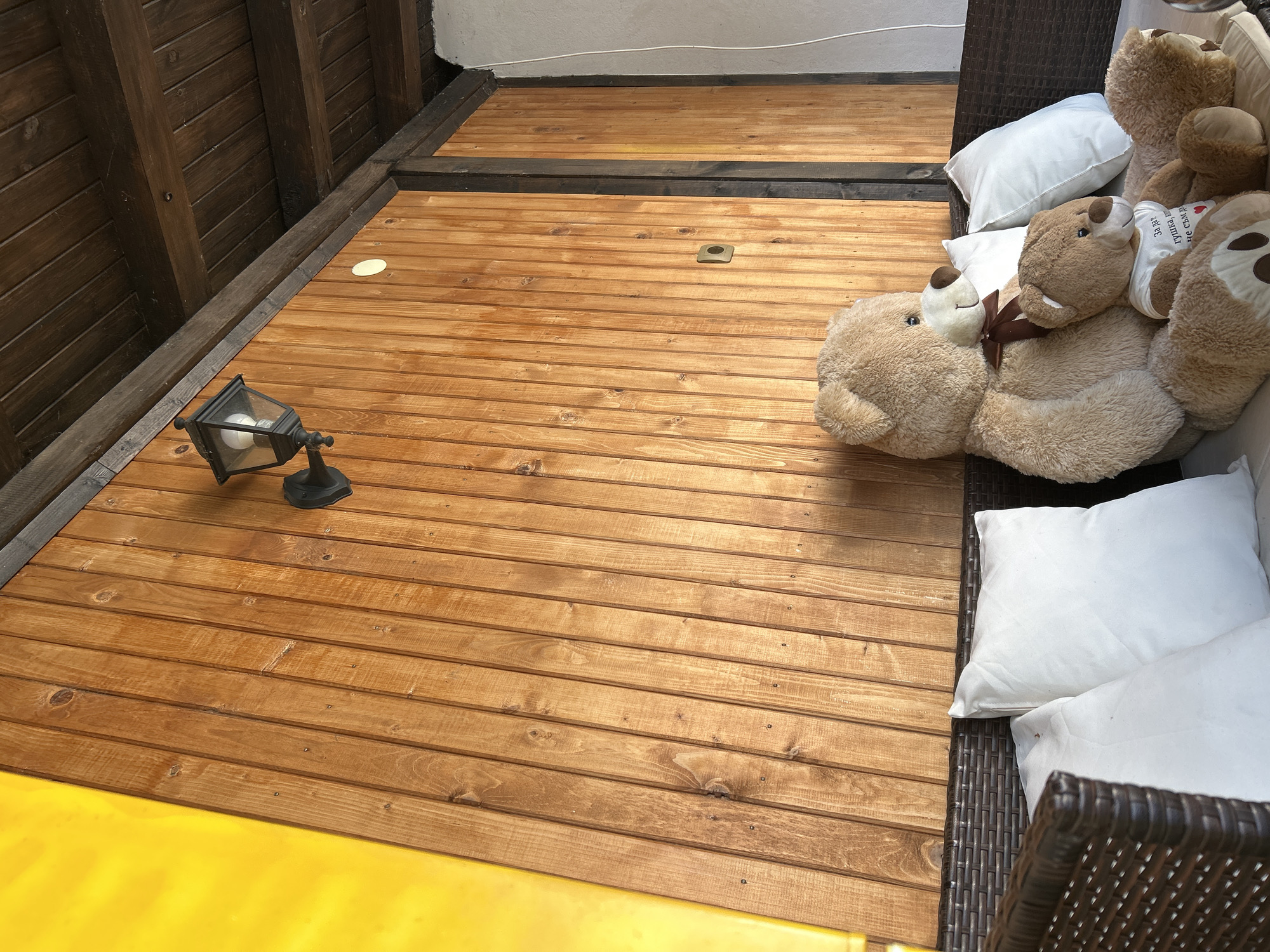 This image captures a cozy, semi-outdoor living space, likely a patio or veranda, characterized by extensive use of wood. The floor consists of light brown, horizontally laid wooden planks, featuring visible wood grain and knots. On the left, a dark brown wooden wall or vertical structure is constructed from vertical planks. The background reveals a lighter section of wooden flooring, separated by a dark horizontal beam, suggesting a multi-tiered structure or different zones. A partial view of a white wall is visible in the upper background.
In the foreground, a black, old-fashioned lantern-style lamp with a clear glass shade and a visible light bulb sits on the wooden floor. Towards the center of the floor, a small square metallic cover and a circular light-colored patch are embedded. The bottom left corner reveals a bright yellow surface, possibly a slide or a colorful mat.
On the right side, dark brown wicker furniture, appearing to be a lounge chair or sofa, is adorned with several white pillows. Resting comfortably on and around these pillows are multiple large, light brown teddy bears. One teddy bear sports a dark brown bow tie, while another wears a white shirt with Cyrillic text. The text on the shirt reads "Я не сплю, я отдыхаю," which translates to "I am not sleeping, I am resting." The scene is brightly lit, indicative of daytime. No human subjects are present, and the arrangement of teddy bears suggests a whimsical or relaxing atmosphere.