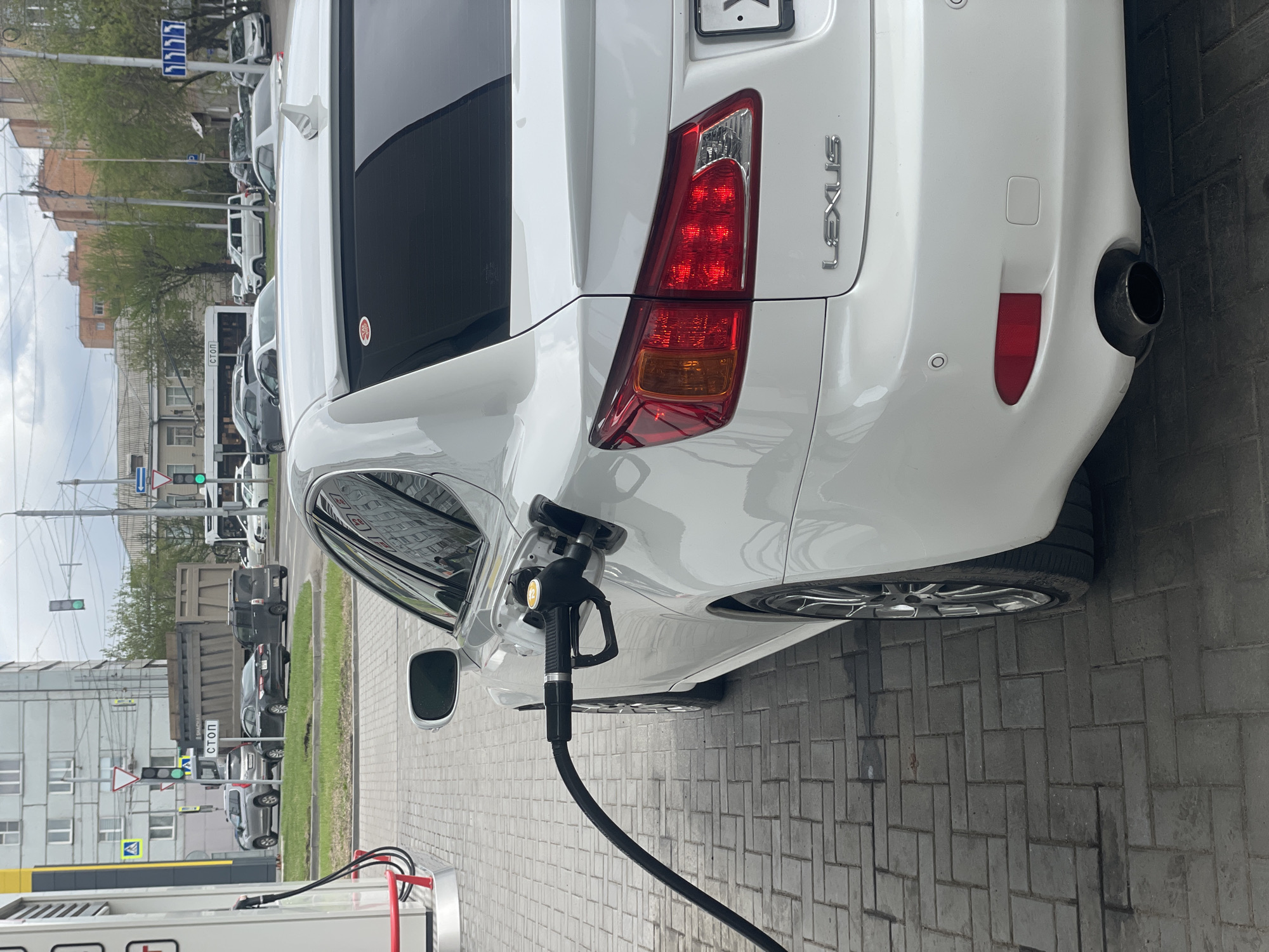 A white Lexus sedan is being refueled at a gas station in Krasnoyarsk, Russia. The car is parked on a brick-paved area near a pump. A black fuel nozzle is inserted into the car's fuel tank. The rear of the vehicle is visible, showing the taillight, exhaust pipe, and Lexus emblem. In the background, buildings, traffic lights, road signs, and other vehicles are visible, indicating an urban environment. A "STOP" sign is visible in Cyrillic lettering. The license plate of the Lexus is partially obscured.