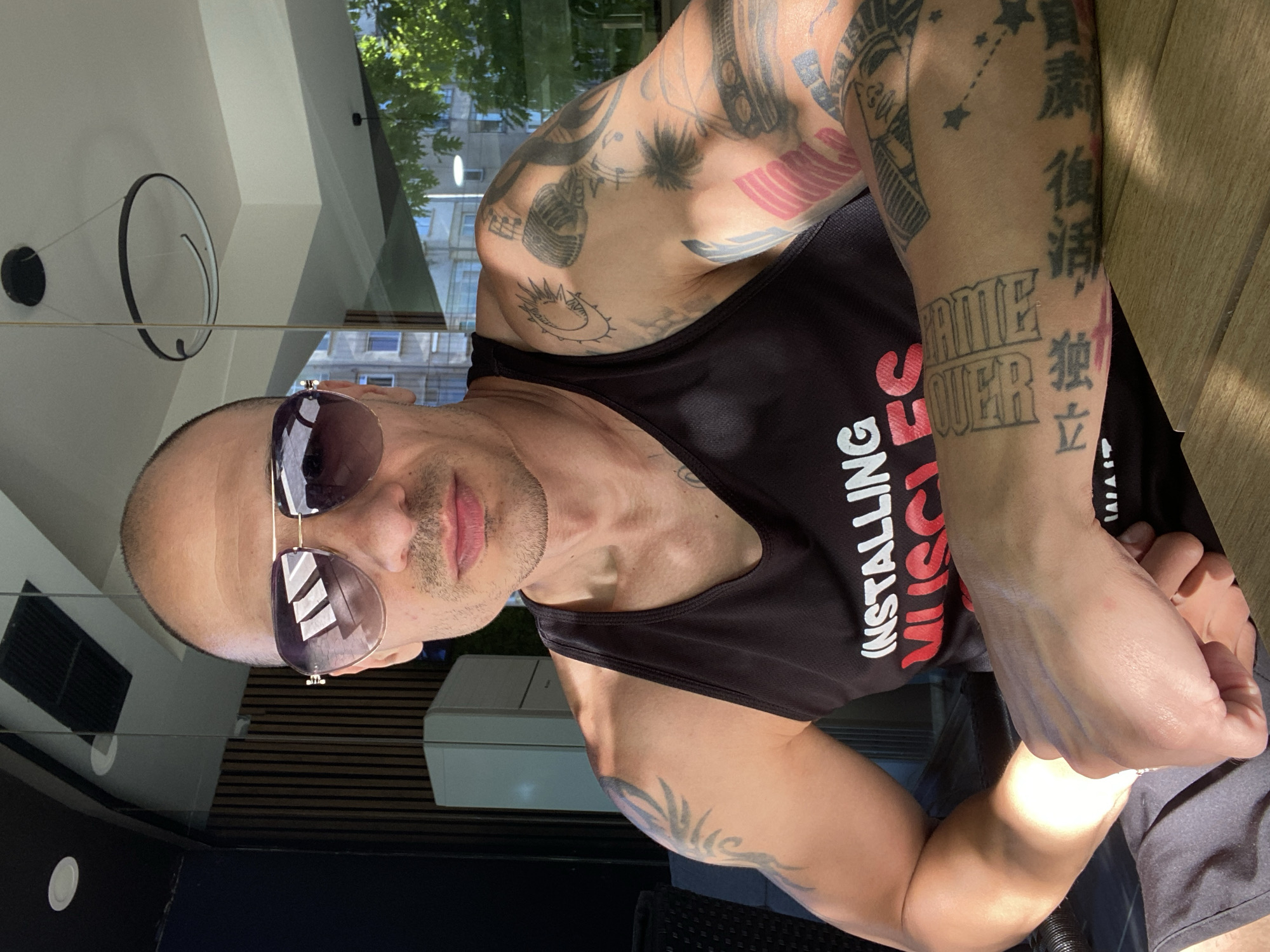 A bald man wearing sunglasses and a black tank top with the words "INSTALLING MUSCLES" in red lettering is sitting at a wooden table outdoors. He has extensive tattoos on his arms. The background shows a modern building with vertical wooden slats and large windows, reflecting the sky and trees. There is a circular light fixture hanging from the ceiling. The man is positioned in the foreground, with his arms resting on the table. This scene is likely located in the city of Pleven, Bulgaria, given the context provided.