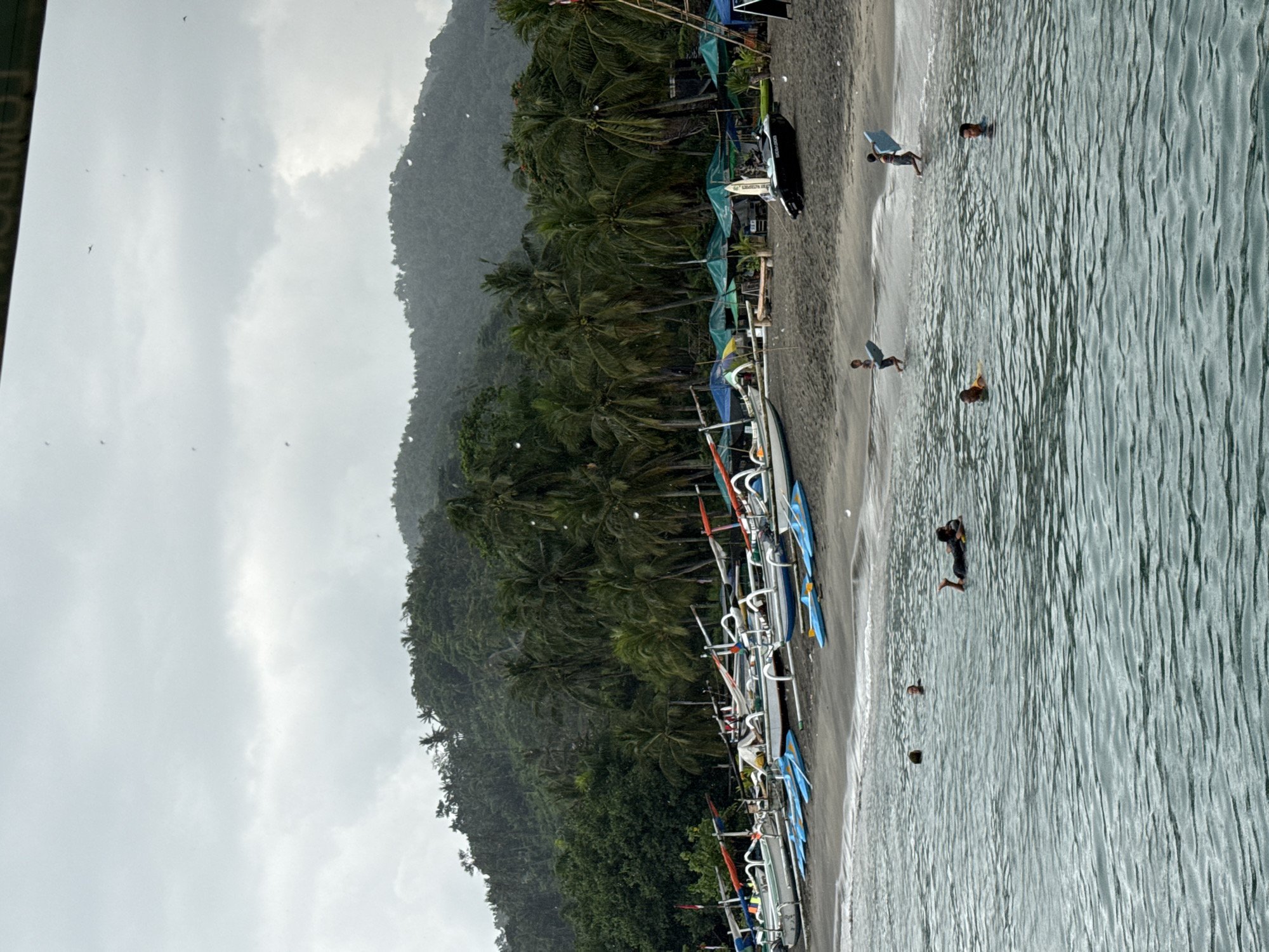 The scene depicts a beach in Senggigi, Indonesia, situated on the coast with boats lining the shore. Lush, green trees and vegetation cover a hill ascending from the shoreline. Above, the sky is overcast. Several people are in the water, some appearing to be surfing or swimming. The water is light blue-green, showing wave patterns.