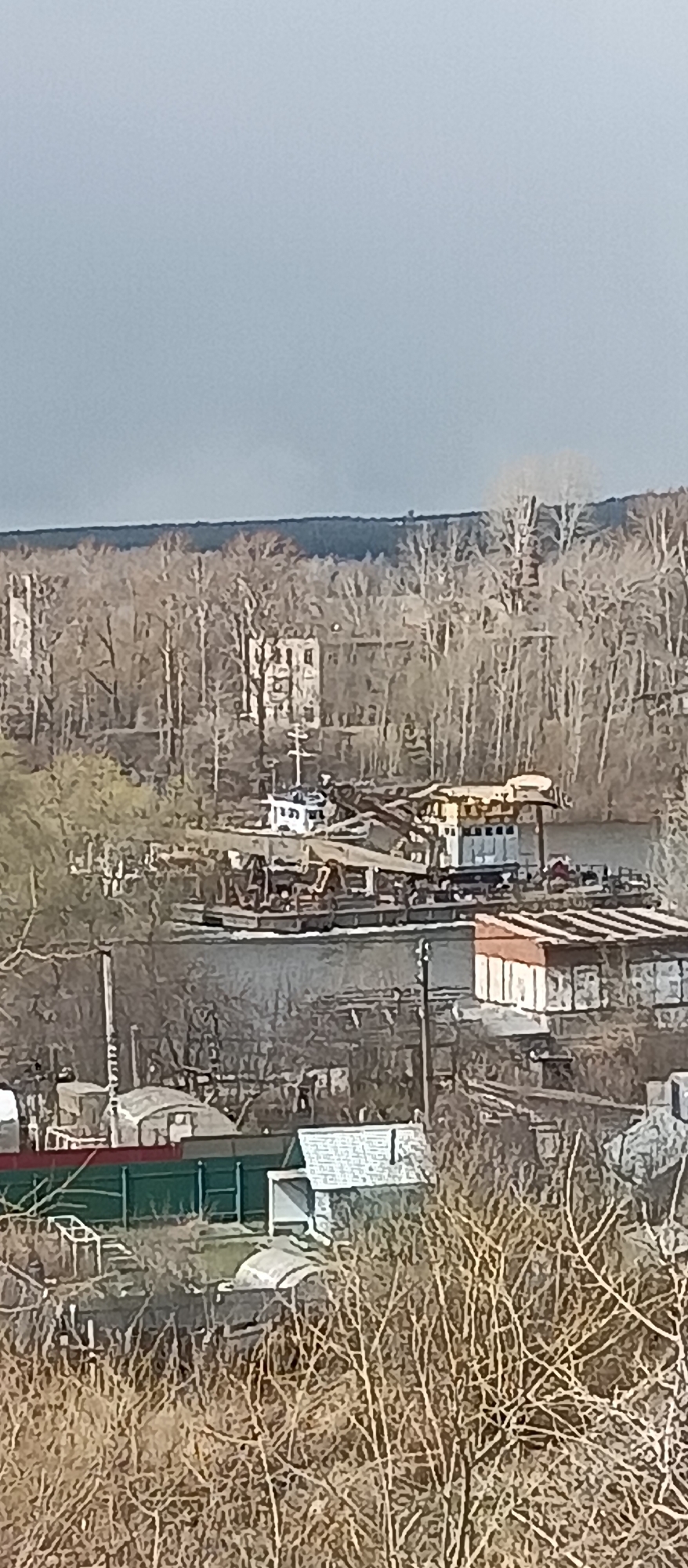 The image presents a wide, elevated view of an outdoor landscape under an overcast, light grey sky. The scene suggests a season between late autumn and early spring due to the prevalence of bare trees and branches.
In the immediate foreground, a dense thicket of leafless branches and dry bushes partially obscures the view. Through these, several rooftops of buildings are visible, including one with a distinct dark green fence and a white building with a patterned roof on the right.
The midground is dominated by a calm body of water, likely a river. On this water, a large, flat-bottomed vessel, possibly a barge or pontoon, is anchored or moving slowly. It carries industrial machinery, appearing to be cranes or excavators. A smaller, white boat is also visible near this larger craft. Along the nearer bank, several low-rise, possibly industrial or storage buildings with reddish-brown roofs are situated.
The background features a vast, dense forest composed entirely of bare, deciduous trees, their intricate branch patterns stretching across the landscape. Interspersed within this forest are several multi-story buildings, their structures and windows discernable among the trees. These buildings appear somewhat weathered or integrated into the natural setting. Beyond this treeline, a faint, darker line of distant trees marks the horizon.
No explicit human activity or individuals are visible, giving the scene a quiet and somewhat desolate ambiance. The industrial equipment on the water suggests ongoing or past work, but no present operation is observed. There is no discernible text anywhere in the image.