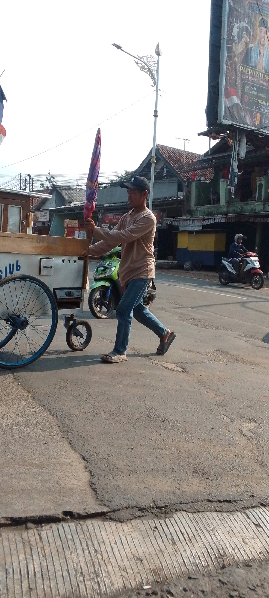 In the city of Cilaku, Indonesia, a man wearing a light brown long-sleeved shirt, blue jeans, and a black baseball cap is walking on the side of a road. He is holding a colorful umbrella with both hands. To his left is a white cart with a wooden top and a large blue-rimmed wheel. A smaller wheel is attached to the cart's front. A green motorcycle is parked behind the cart. In the background, there are several buildings, including one with a large billboard advertising an event featuring a man with a beard and headdress. A white motorcycle with a rider wearing a helmet is visible further down the road. The sky is bright, suggesting daytime. The road surface appears to be asphalt and is worn in places.
