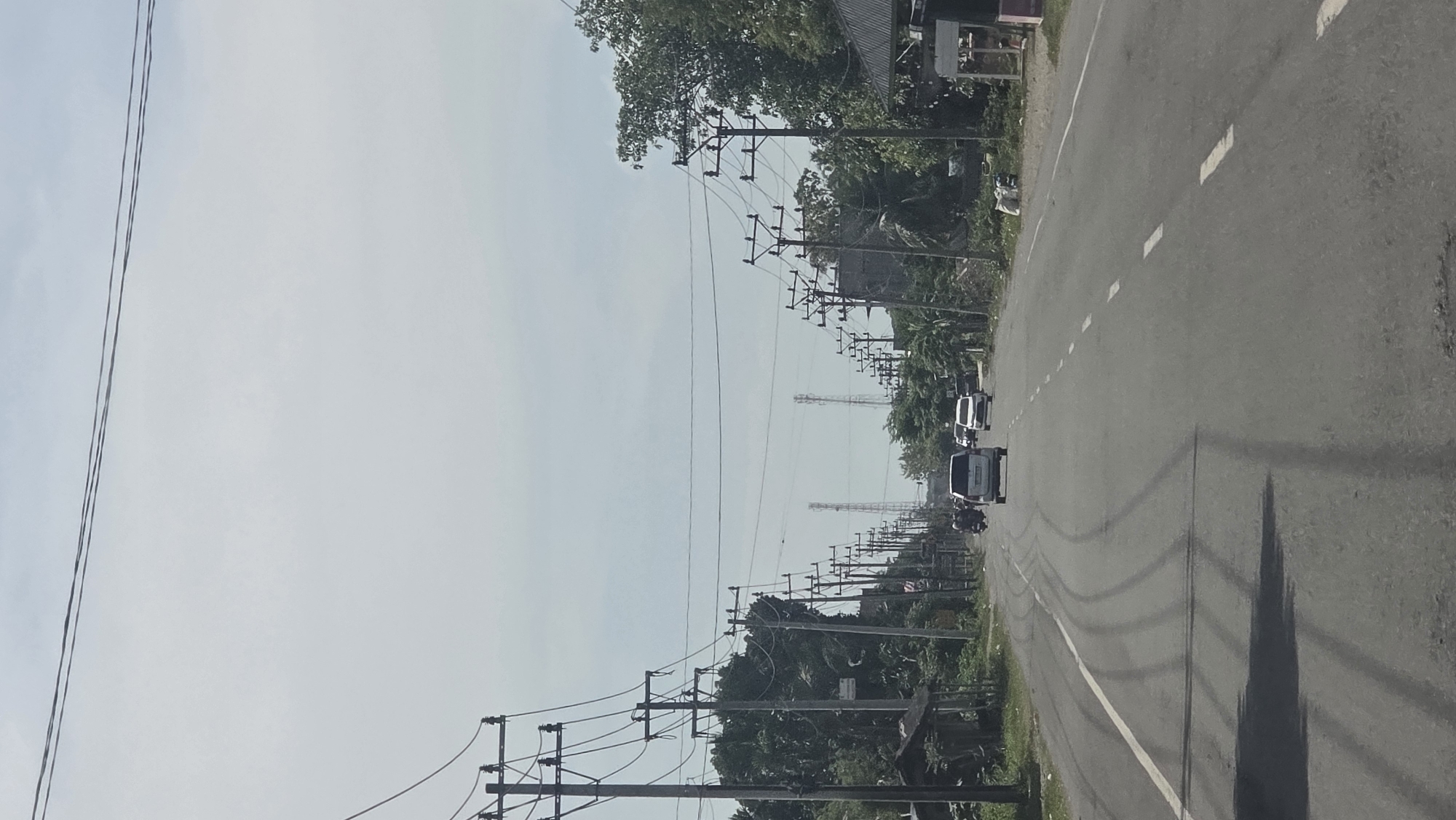 A road scene, likely in Teungoh Baro, Indonesia. The road extends into the distance with a line of utility poles on either side. There are several vehicles on the road, including vans and a motorcycle. The sky is overcast. The road is marked with dashed white lines. Shadows suggest the time of day. Vegetation is visible along the road.