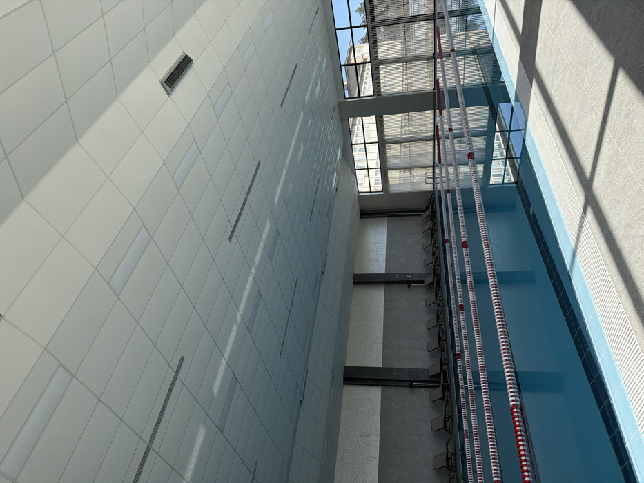 This image depicts the interior of a modern, multi-lane indoor swimming pool facility in Dzhebel, Bulgaria. The large rectangular pool, filled with clear blue water, features at least four lanes demarcated by red and white floating lane lines. The pool deck is tiled with light grey squares, illuminated by natural light entering through a wall of tall, segmented windows on the right. Strong sunlight casts defined shadows onto the deck.
Along the left-hand wall, a row of approximately twelve dark-colored lounge chairs is neatly arranged on a tiled surface adjacent to the pool. The lower portion of this wall is dark grey, while the upper section is a lighter textured grey; dark vertical columns are interspersed along its length. The ceiling consists of a grid of white modular panels, integrating numerous rectangular LED light fixtures, some of which are illuminated, and several dark rectangular ventilation grilles.
Beyond the expansive windows, parts of a multi-story building with numerous windows are visible, suggesting an urban or institutional setting. There are no individuals present in the scene, and no actions or interactions are occurring. The environment appears clean, well-maintained, and unoccupied.