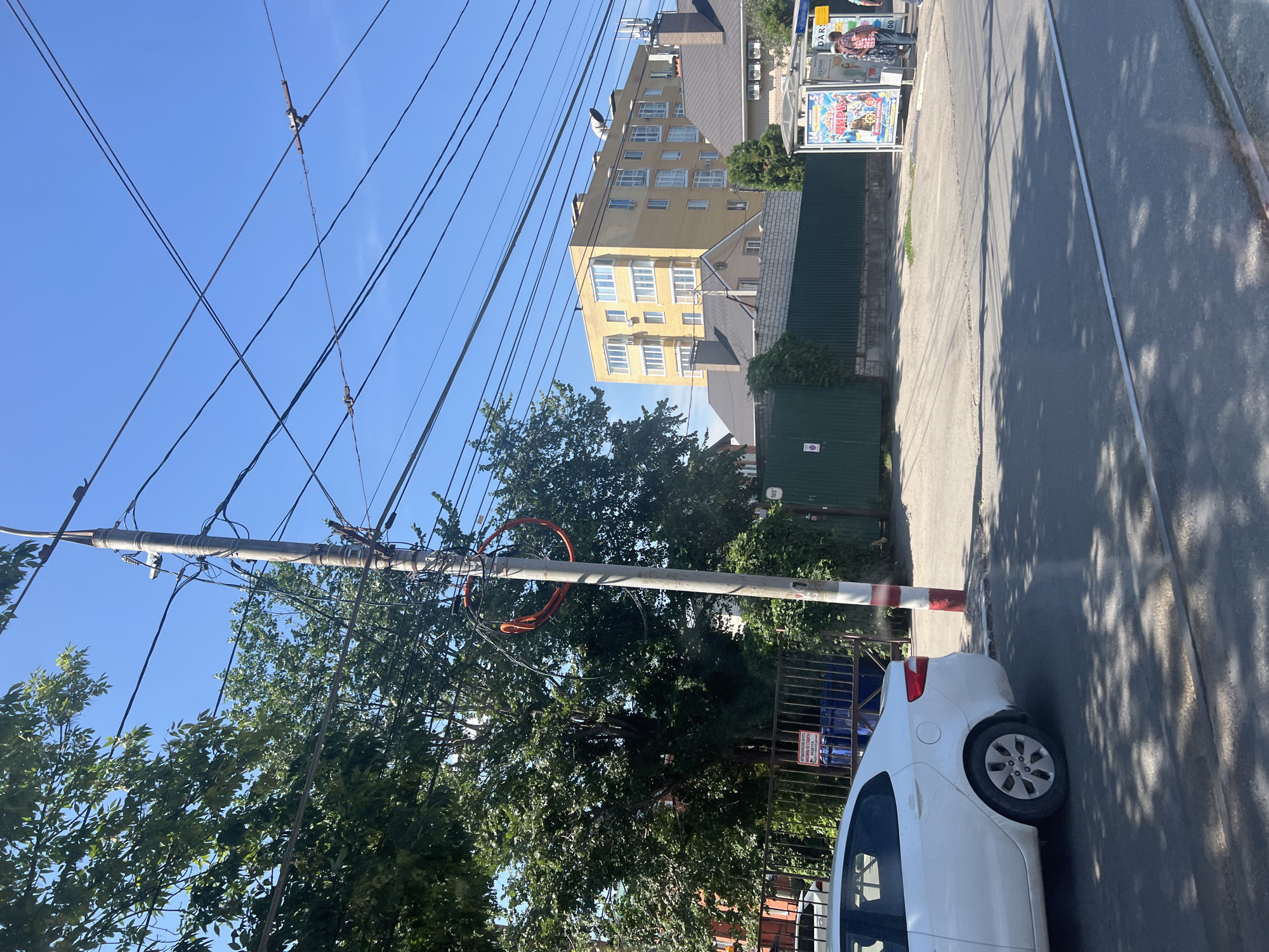 In Ulyanovsk, Russia, on a sunny day, a white sedan is parked near a utility pole adorned with numerous wires and a circular orange object. Behind the pole, a metal fence encloses a blue dumpster, with a sign in Russian attached to the fence. Beyond the fence, a tall, yellow apartment building stands against a clear blue sky. To the right, a bus stop shelter displays advertisements, and two individuals are standing nearby. The scene is a street with a paved road and trolley tracks visible.