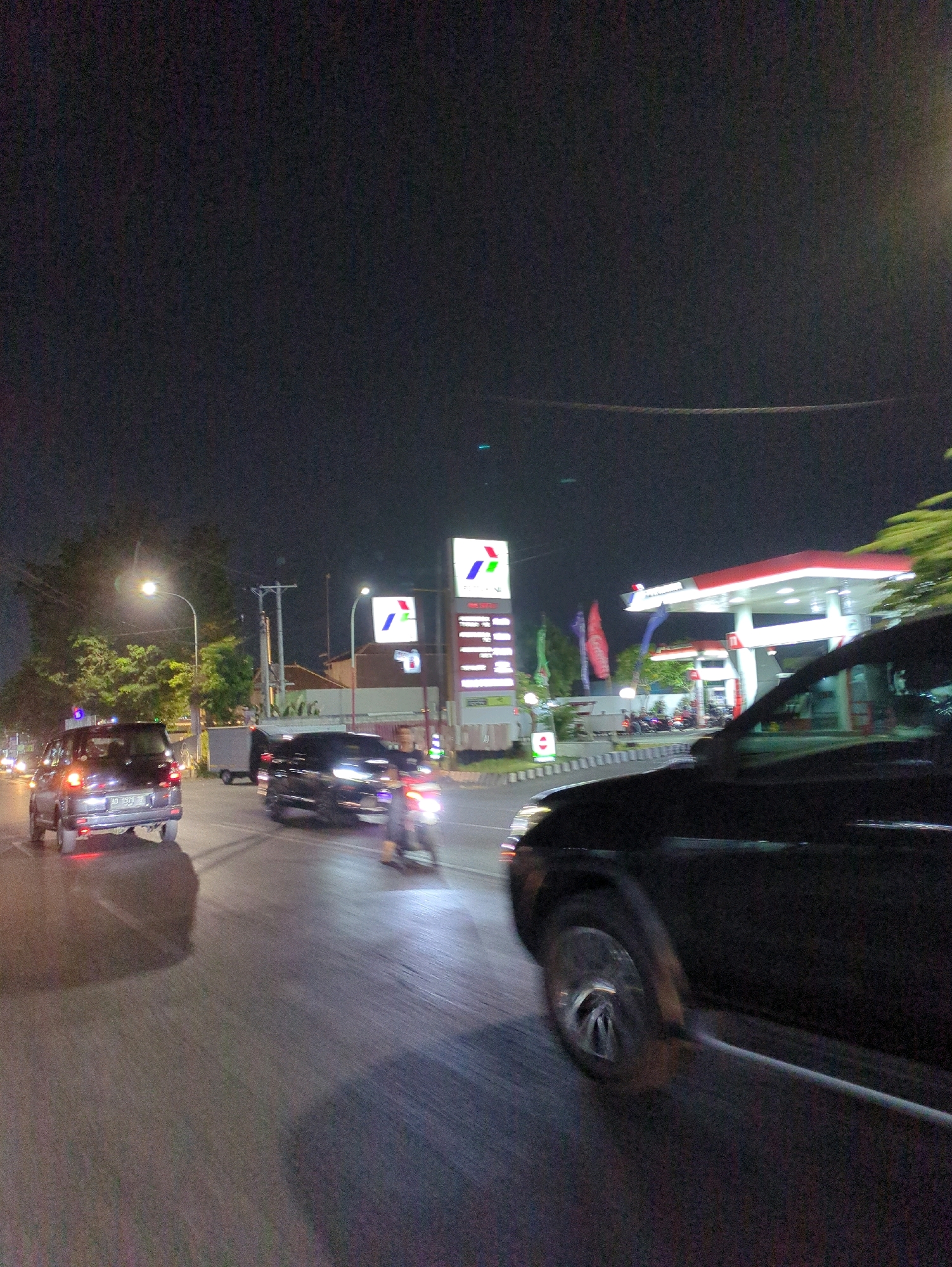 The image depicts a night scene at a gas station in Malangjiwan, Indonesia. A black SUV is prominently featured in the foreground, angled towards the right of the frame. To the left, a silver SUV is visible with its taillights illuminated. Several other vehicles, including a motorcycle, are in motion on the road, creating motion blur. The gas station itself is illuminated by bright lights, with a large red and white canopy. Signage with the Pertamina logo is visible. Trees and streetlights are also present in the environment. The overall impression is one of activity on a street at night, with a focus on vehicles and the gas station as a landmark.