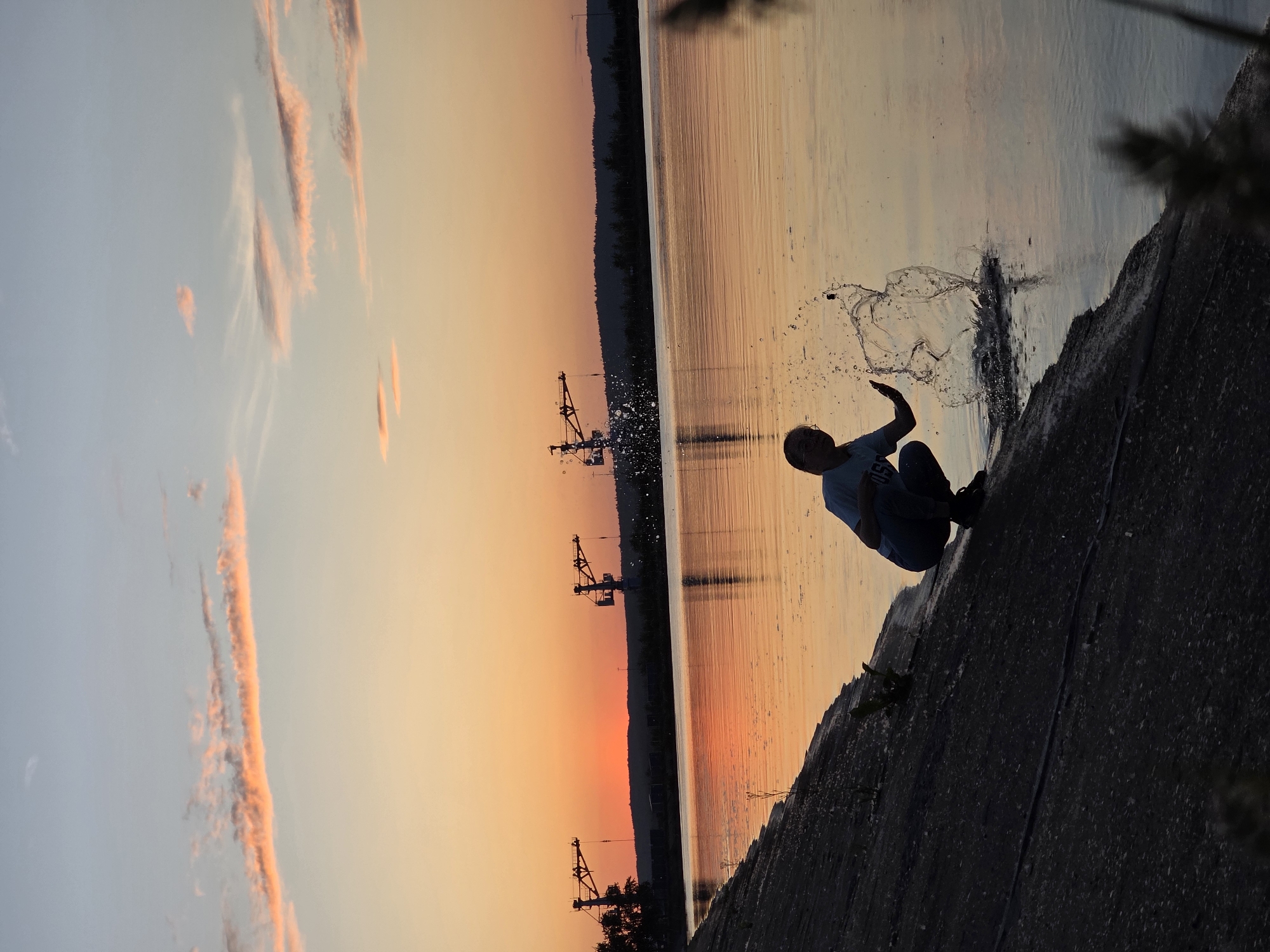 The image captures a sunset scene in Chaikovsky, Russia, along what appears to be the Kama River or Reservoir. In the foreground, a silhouetted individual is crouched on a dark, sloping concrete embankment at the water's edge. The person, visible wearing glasses and a light-colored shirt, is actively splashing the water with their hand, creating a significant, multi-peaked splash of clear water droplets and larger liquid masses. The wide body of water in the midground reflects the vibrant hues of the setting sun. The sky above transitions from an intense orange and red directly at the horizon to a lighter, soft blue overhead, featuring scattered, elongated clouds tinged with orange and pink. In the distant background, several large industrial cranes are silhouetted against the bright horizon, positioned over a dark, tree-lined landform, consistent with the industrial activity around the Votkinsk Hydroelectric Power Plant. The overall scene is backlit, casting the foreground and the person into deep shadow while highlighting the colorful sky and its reflection on the water.