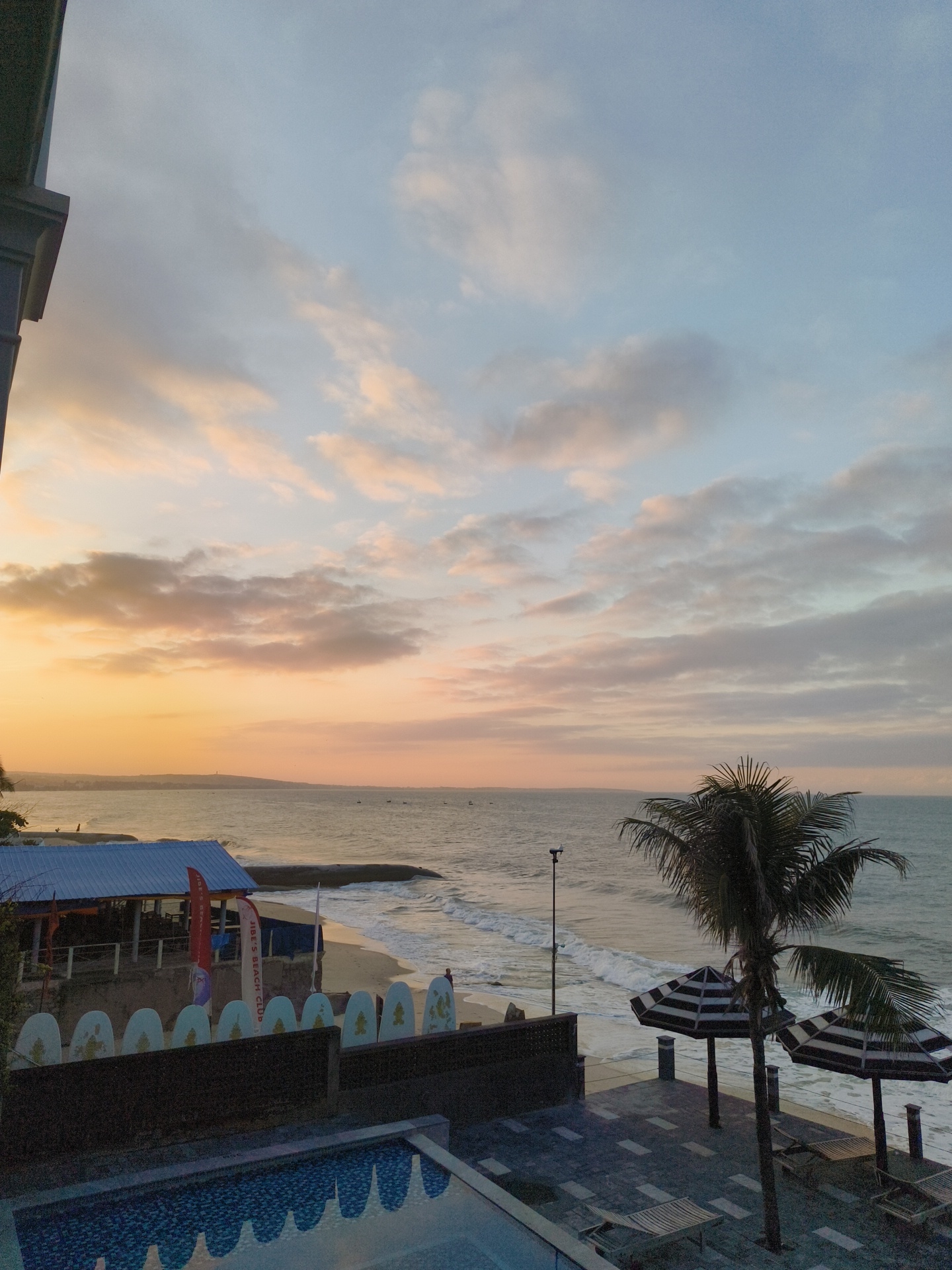 This media file depicts a serene beach scene at sunset in Phu Thuy Ward, Vietnam. The sky is filled with a blend of soft orange, pink, and pale blue hues, indicating the twilight hours. Wispy clouds are scattered across the sky, catching the last rays of the setting sun.
The foreground features a swimming pool with a patterned tiled bottom, reflecting the sky. Adjacent to the pool is a paved patio area with lounge chairs and two striped umbrellas, suggesting a resort or hotel setting. A lone palm tree with its fronds swaying in the gentle breeze stands tall near the umbrellas, adding a tropical ambiance.
Beyond the patio, a sandy beach meets the ocean. Waves are gently rolling onto the shore. On the beach, there's a small building with a blue roof, possibly a beach bar or cafe, adorned with flags. A line of surfboards, white with gold accents, are propped up against a dark wall, hinting at a surf club or rental. A few small boats are visible on the horizon.
There is a subtle feeling of tranquility and relaxation, characteristic of a beach at dusk. The limited visibility of people suggests a quiet, perhaps end-of-day atmosphere. The text "JOE'S BEACH CLUB" is visible on one of the flags. The overall scene is picturesque and calming, capturing the beauty of a coastal sunset.