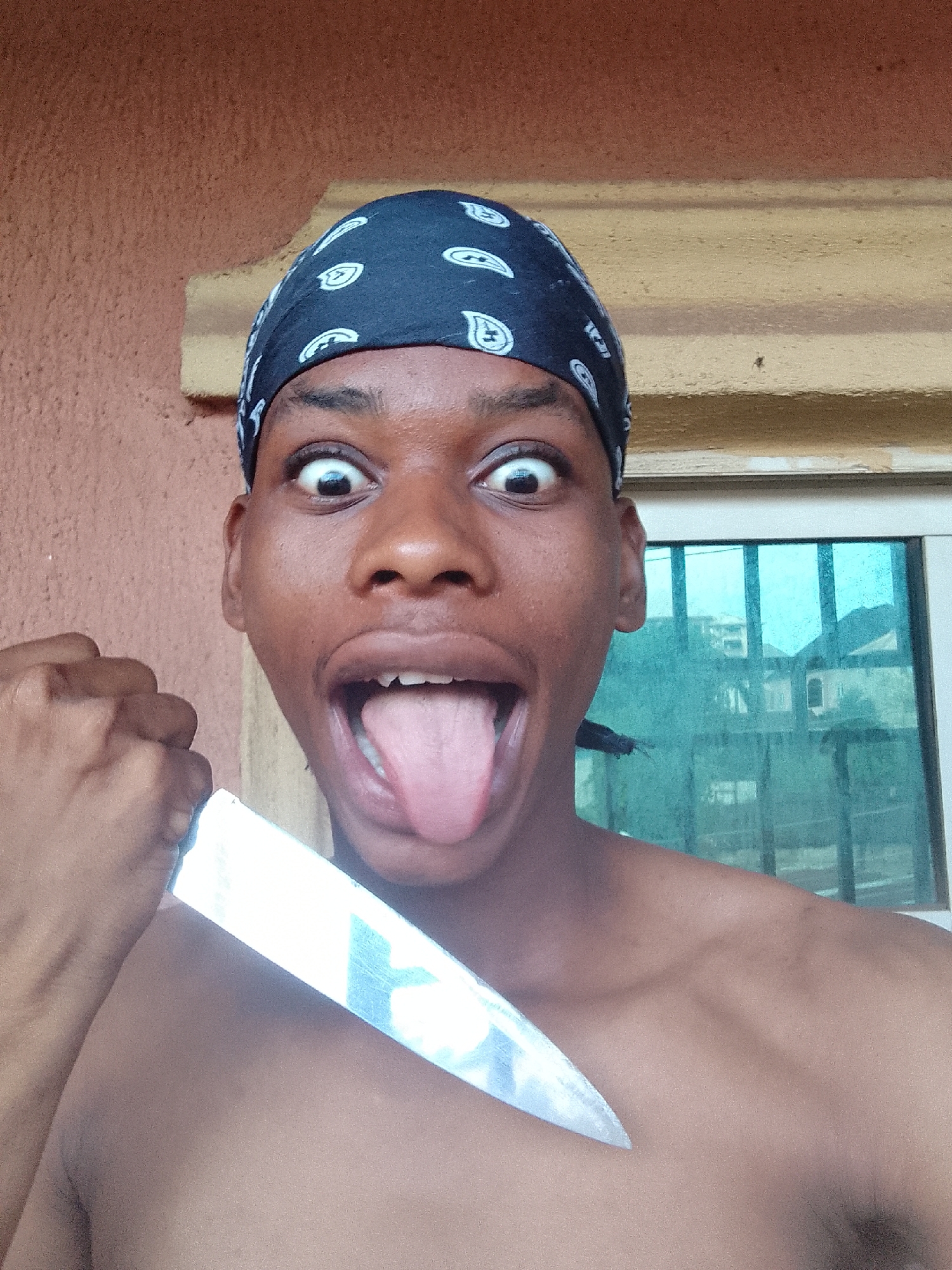 A shirtless person with brown skin is holding a large knife near their chest and making a face with their eyes wide open and tongue sticking out. The person is wearing a black bandana with white paisley designs. Behind them is a window with blue-tinted glass and white bars. Outside the window are buildings, and the background is out of focus. The wall behind the person is a light reddish-brown. This scene may be in Onitsha, Nigeria due to the prompt.
