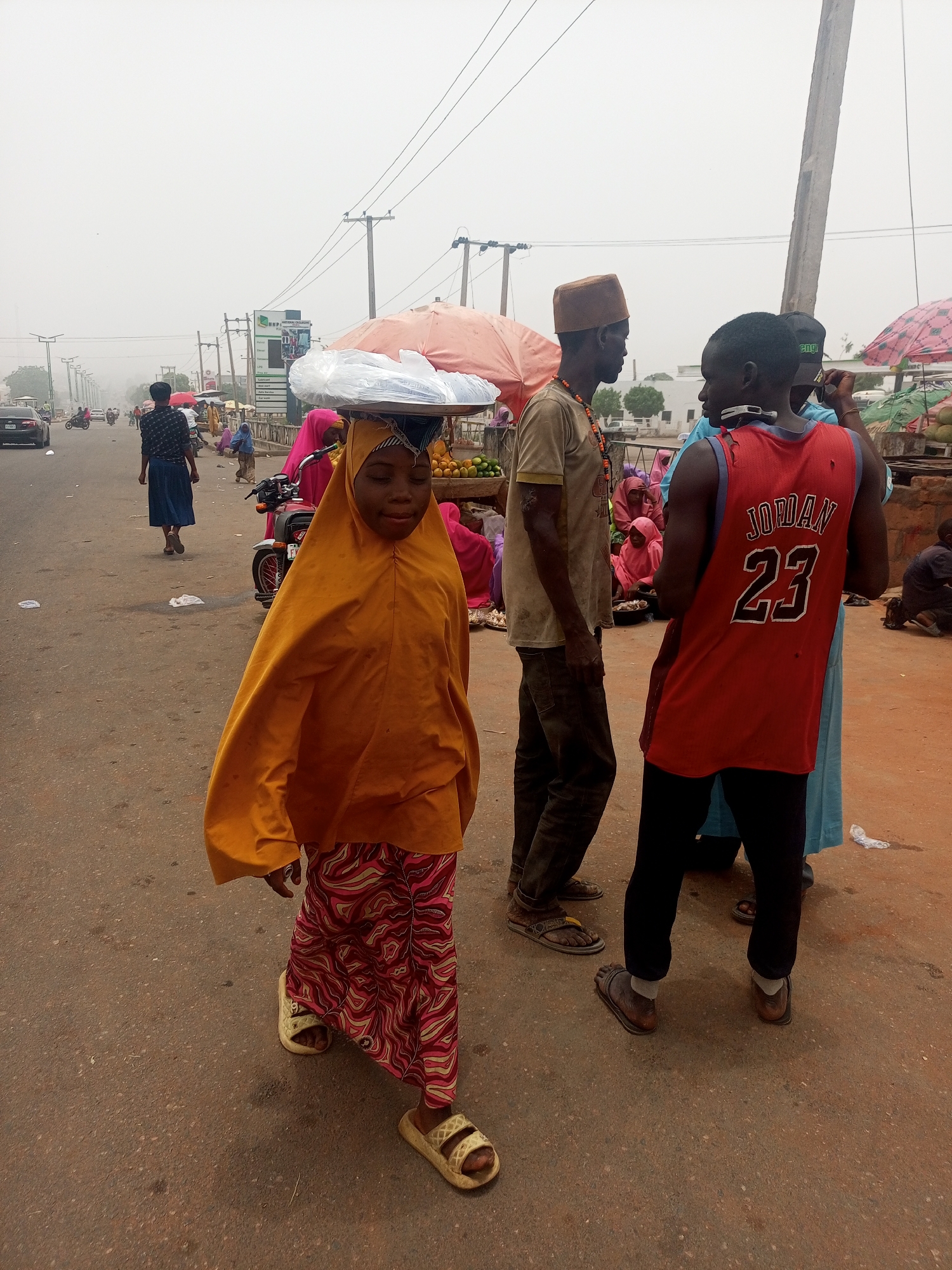 This outdoor photograph captures a vibrant street scene, likely a market area, in Tempure, Nigeria, under an overcast or hazy sky. The central figure is a young person, possibly female, walking towards the viewer, expertly balancing a flat tray piled with plastic-wrapped items on her head. She is dressed in a flowing yellow head covering (hijab or similar) and a patterned skirt in shades of pink, red, and white, wearing light-colored slide sandals.
To the right of the frame, a man in a brown traditional cap and a light-colored t-shirt stands, facing left. Beside him, another man is seen from behind, wearing a red sleeveless basketball jersey with "JORDAN 23" printed in white on the back. He wears dark pants and sandals.
The background is filled with activity, showing other individuals walking, congregating, and tending to roadside stalls. Two prominent market umbrellas, one pinkish-red and another orange, provide shade over visible goods such as fruits and produce. A motorcycle is parked nearby. In the distance, a busy road stretches, with several cars and motorcycles indicating ongoing traffic. Utility poles and power lines are visible against the hazy sky. A green and white signboard with the text "NATIONAL EXCLUSIVE" is partially visible in the mid-ground. The scene depicts everyday life and commerce in a bustling community.