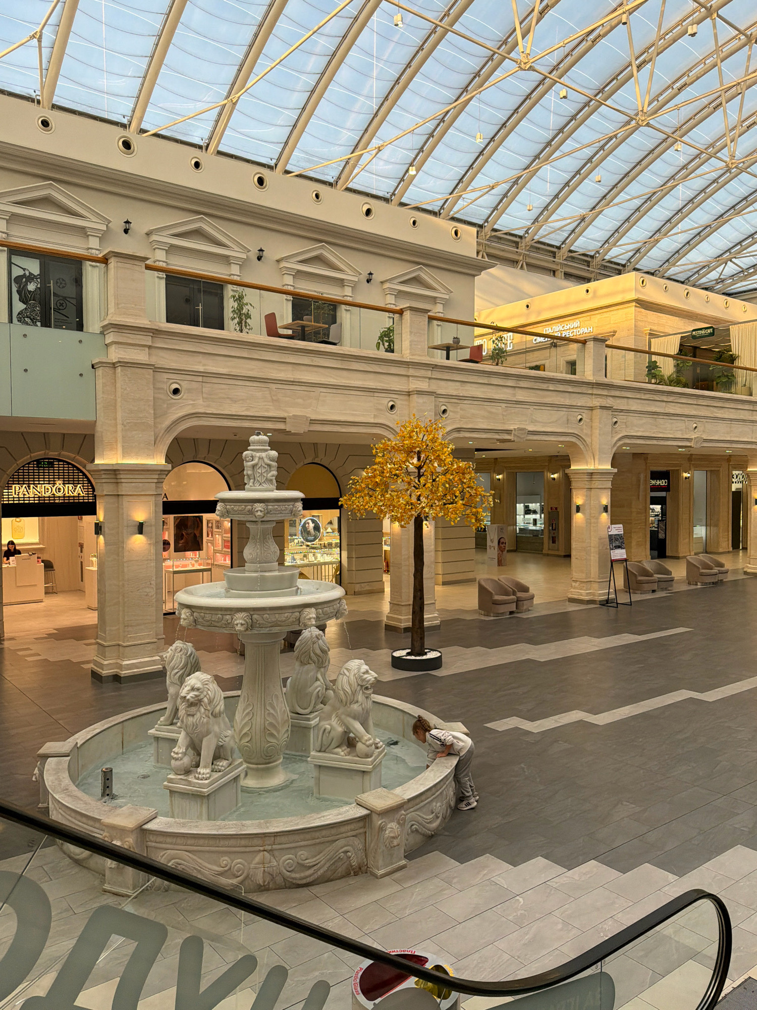 This is a photograph taken inside a modern shopping mall. The mall features a large, open atrium with a glass roof that allows natural light to filter in. Several floors are visible, connected by walkways and balustrades. A prominent white marble fountain with lion sculptures is the central focal point in the foreground. A young child is seen leaning over the edge of the fountain, looking into the water. To the left of the fountain is the entrance to a Pandora jewelry store. To the right, a decorative artificial tree with yellow foliage stands near a row of seating areas. The floor is a tiled surface with a patterned design. In the background, shop fronts and signage are visible, indicating a variety of retail establishments. The overall ambiance suggests a well-maintained and upscale commercial environment. The location is the Dream Town shopping mall in Kyiv, Ukraine.