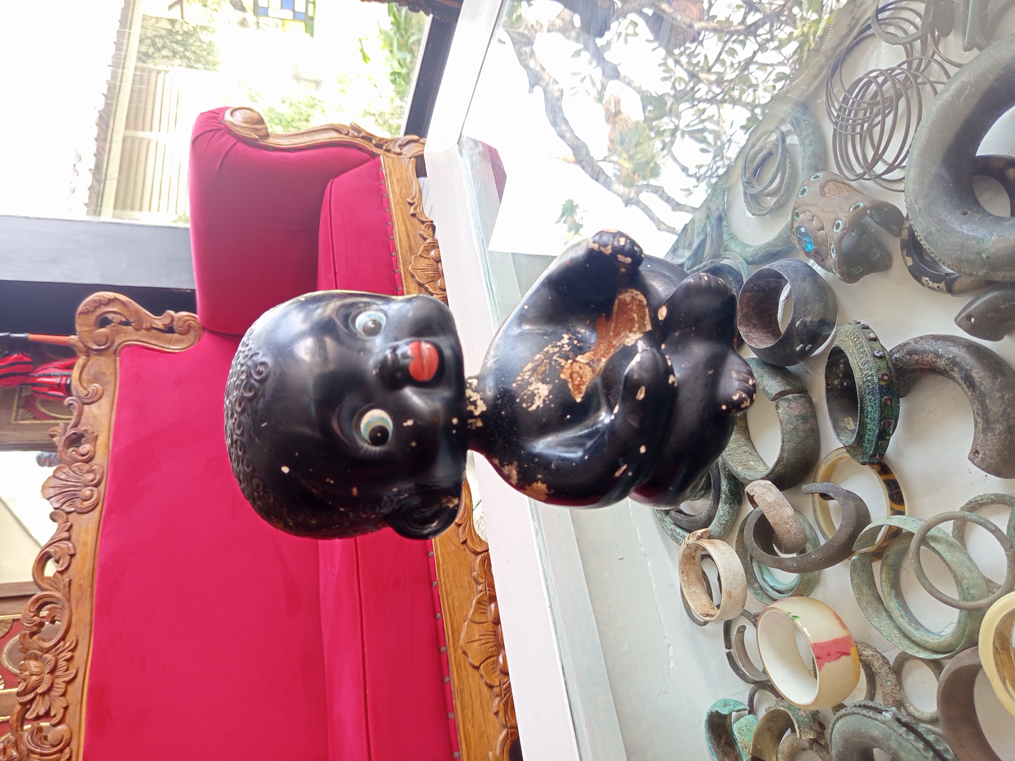 A figurine of a black baby with its tongue sticking out is positioned in front of a display case containing various bracelets and rings. The figurine is situated near a red chair with ornate wooden detailing. The scene appears to be in North Kuta, Indonesia.