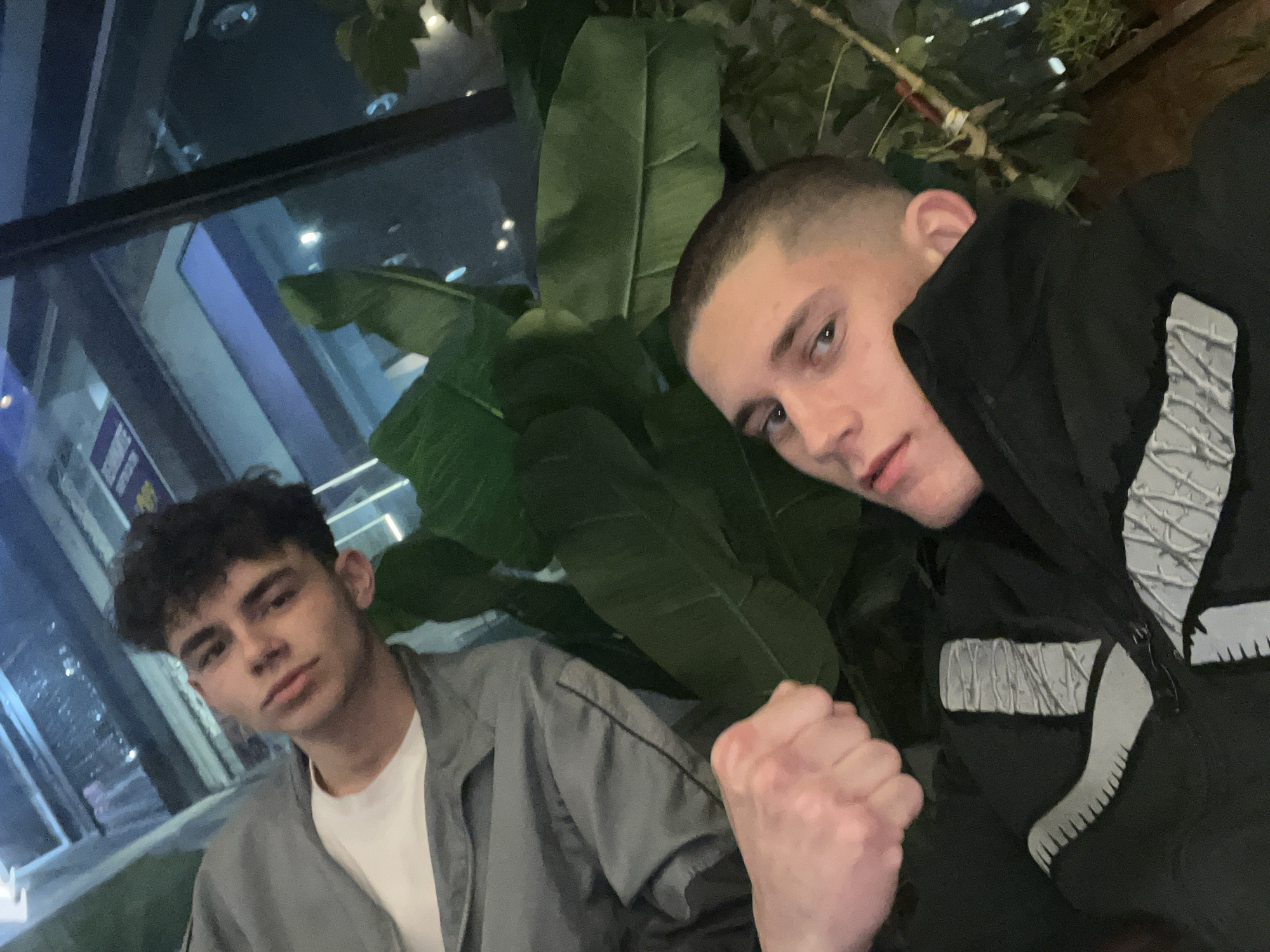 Two young men pose for a selfie in an indoor setting with greenery. One man has short dark curly hair, wearing a white t-shirt and grey jacket. The other has a very short buzzcut and is wearing a black jacket with white designs. They are indoors, near a window with city lights and buildings visible outside, possibly indicating the city of Tsarevo, Bulgaria. The presence of the large leaves suggests the location might be a restaurant or indoor space with plants for decoration.