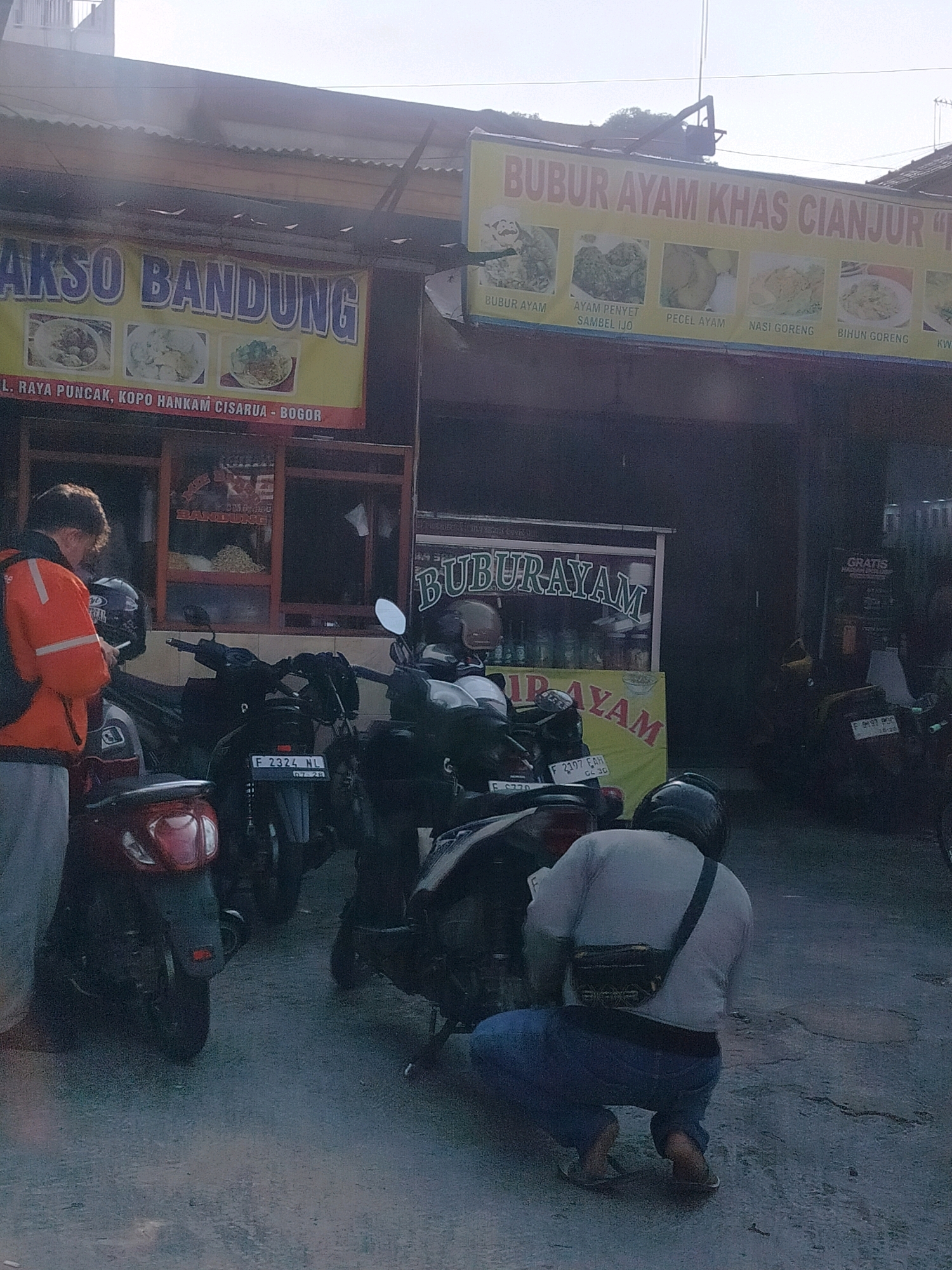 The image depicts a street scene in Cisarua, Indonesia, likely a commercial area with food vendors. A yellow banner with red lettering reading "AKSO BANDUNG" is visible, featuring images of food. Below this banner, another yellow banner displays "BUBUR AYAM KHAS CIANJUR" (Cianjur Specialty Chicken Porridge) with images of various dishes such as bubur ayam, ayam penyet, pecel ayam, nasi goreng, and bihun goreng. A portion of a stall with the name "BANDUNG" is visible behind the man in orange.
In the foreground, a man wearing an orange jacket with reflective strips and a black backpack is looking at his phone. He is standing next to a red scooter. Behind him, several motorcycles are parked. Further into the scene, another man wearing a light-colored shirt, dark pants, and a helmet is crouching down, looking towards the ground. He has a fanny pack worn across his chest. Motorcycles are also parked behind him. A sign with "GRATIS HADIAH LANGSUNG" (Free Instant Prize) is visible on the right side of the image. The ground appears to be asphalt, with some wet patches and cracks. The overall environment suggests a busy outdoor food stall area.
