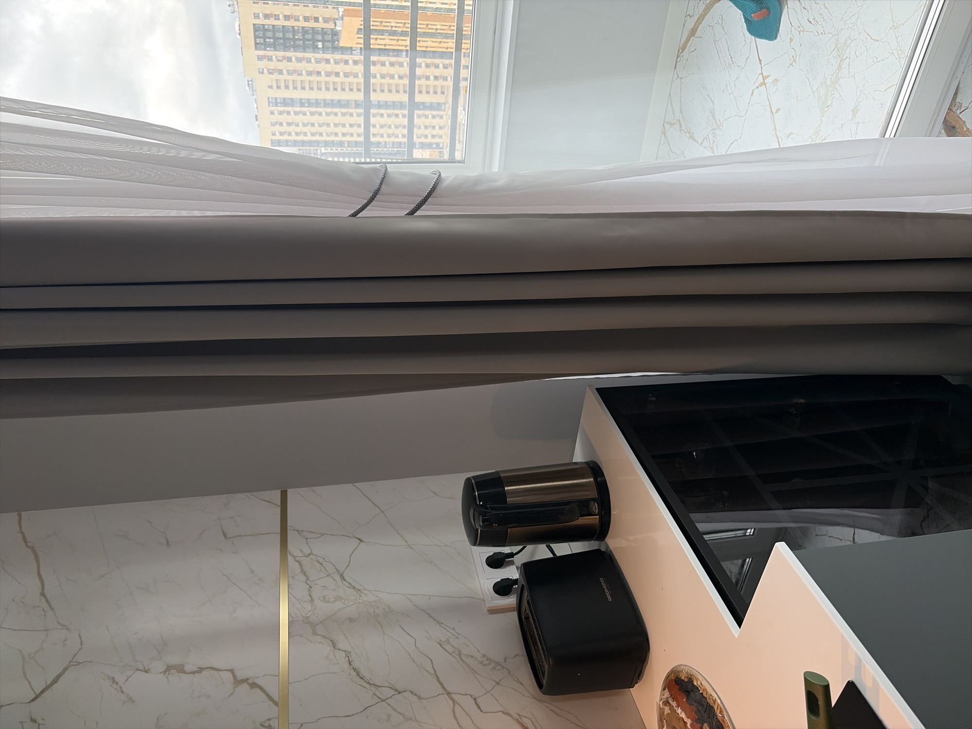 This is a medium shot, indoors, of a kitchen counter with appliances and a window with curtains. The counter is white and appears to be a laminate or quartz material. On the counter are a black toaster, a silver and black electric kettle, and a small plate with some food items. Behind the counter is a wall with a marble-like pattern and a horizontal gold trim. To the right of the counter are two electrical outlets with plugs inserted.
The kitchen area is adjacent to a large window covered by two layers of curtains. The outer curtain is a thick, gray fabric, and it is held back with a decorative rope or chain tie. The inner curtain is sheer white. Through the window, a cityscape is visible, with apartment buildings in the background. The sky is overcast. The floor visible in the foreground and through the window also has a marble pattern. The overall impression is of a modern, well-lit kitchen.
There are no people visible in the image. The scene appears to be a still life of kitchen appliances and decor. Based on the architectural style of the buildings visible through the window, the image could be from a modern residential area in Moscow, Russia.