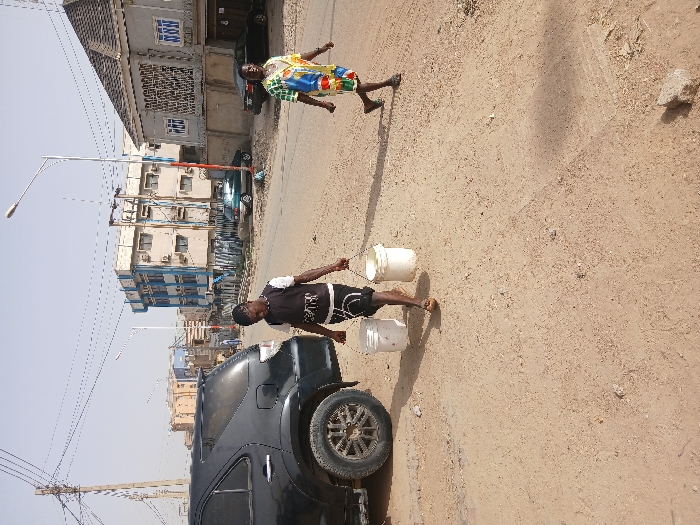 The scene depicts an outdoor street environment in Mgbidi, Nigeria, under clear daylight conditions. Two individuals are walking on a dirt shoulder adjacent to a paved road.

In the foreground-center, a male subject walks from right to left, wearing a black sleeveless shirt with white graphics and black shorts, along with flip-flops. He carries two white plastic buckets, suspended by wire handles, one in each hand. His attention is directed forward.

In the mid-ground, on the right side of the frame, a female subject walks from left to right. She wears a brightly colored, patterned, loose-fitting dress and flip-flops. Her gaze is directed forward.

On the far left, a black SUV, possibly a Toyota Fortuner, is parked on the dirt shoulder, with its rear and right side visible. Tire tracks are evident in the light brown, sandy dirt foreground. A paved road occupies the mid-ground, with a light blue sedan parked further down on the right side of the road. Another dark-colored sedan is parked behind the walking female.

In the background, a multi-story building with blue trim and multiple windows/air conditioning units is prominent on the left. To the right, another building with a grey tiled roof and decorative window grilles is visible. A network of overhead utility lines and several utility poles, including at least two streetlights, are present against the clear sky. Shadows are cast by the individuals and vehicles, indicating direct sunlight.