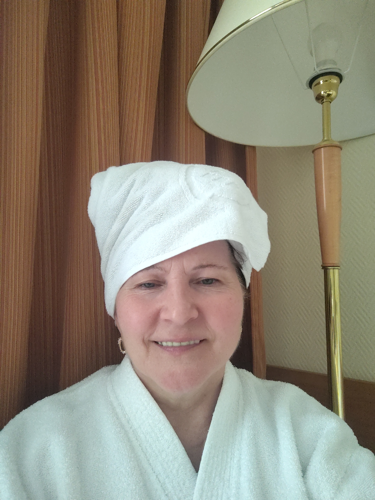 A middle-aged woman is the central subject of this close-up, chest-up portrait, captured indoors in what appears to be a private setting, likely a hotel room or bathroom in Moscow, Russia. She has a white terry cloth towel wrapped around her head, suggesting she has recently washed her hair, and is wearing a matching white terry cloth bathrobe. Her face is turned slightly towards the viewer, with a gentle, relaxed smile and crinkled eyes indicating contentment. A small, decorative hoop earring is visible on her right ear.
The background features warm tones and textures. To the left, there are vertical-striped curtains in shades of brown and orange. To the right, a cream-colored lampshade with a golden rim is prominent, along with the wooden and brass stem of a lamp. Behind her, a light beige wall with a subtle, geometric pattern is visible. The lighting is soft and even, typical of indoor conditions. There is no visible text in the image. The scene conveys a sense of comfort and relaxation, possibly after a refreshing bath or shower.