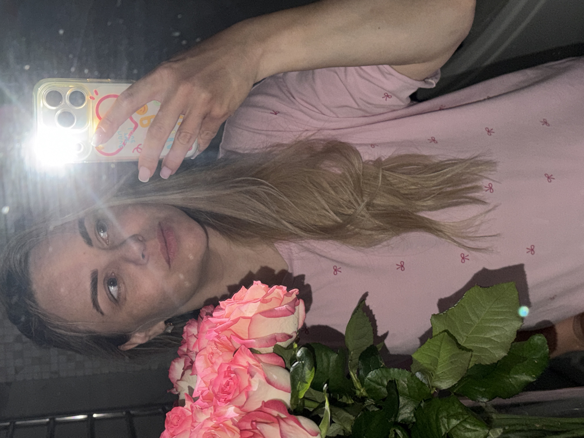 A woman is holding a bouquet of pink roses and taking a selfie with her smartphone. The smartphone flash is on. The woman is wearing a pink shirt with a bow pattern. Her hair is blonde. The location cannot be determined to be Votkinsk, Russia, as no location-specific details are present in the image.