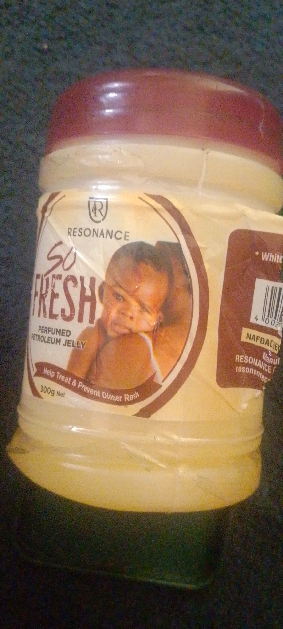 This media file shows a close-up shot of a jar of "So Fresh" perfumed petroleum jelly. The jar has a light yellow base and a reddish-brown lid. The label on the jar features a circular image of a baby's face, with the text "RESONANCE" and a logo with the letter "R" inside a shield. Below the baby's image, the label further states "PERFUMED PETROLEUM JELLY" and "Help Treat & Prevent Diaper Rash." The net weight of the product is indicated as "300g Net."
To the right of the main label, there is a smaller brown label with a barcode and the text "White." Below this, partially visible, is "NAFDACIED" and "RESONANCE / Manul / resonantec." The jar appears to be placed on a dark, textured surface, possibly carpet or fabric. The lighting is somewhat dim, and the image has a grainy quality, suggesting it might have been taken with a low-light setting or older camera. No specific time of day, weather, or discernible location cues are present beyond the product itself. The focus is on the product, with the background out of focus.