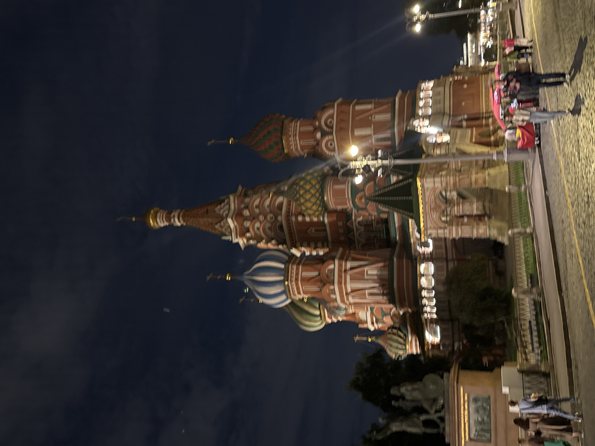The image captures Saint Basil's Cathedral in Moscow, Russia at night. The iconic onion domes and intricate architecture of the cathedral are illuminated, standing out against the dark, cloudy sky. Several people are visible walking on the cobblestone street in front of the cathedral, some are gathered around stalls with red umbrellas. Streetlights cast a warm glow on the scene. The overall atmosphere is that of a bustling tourist area at night.
