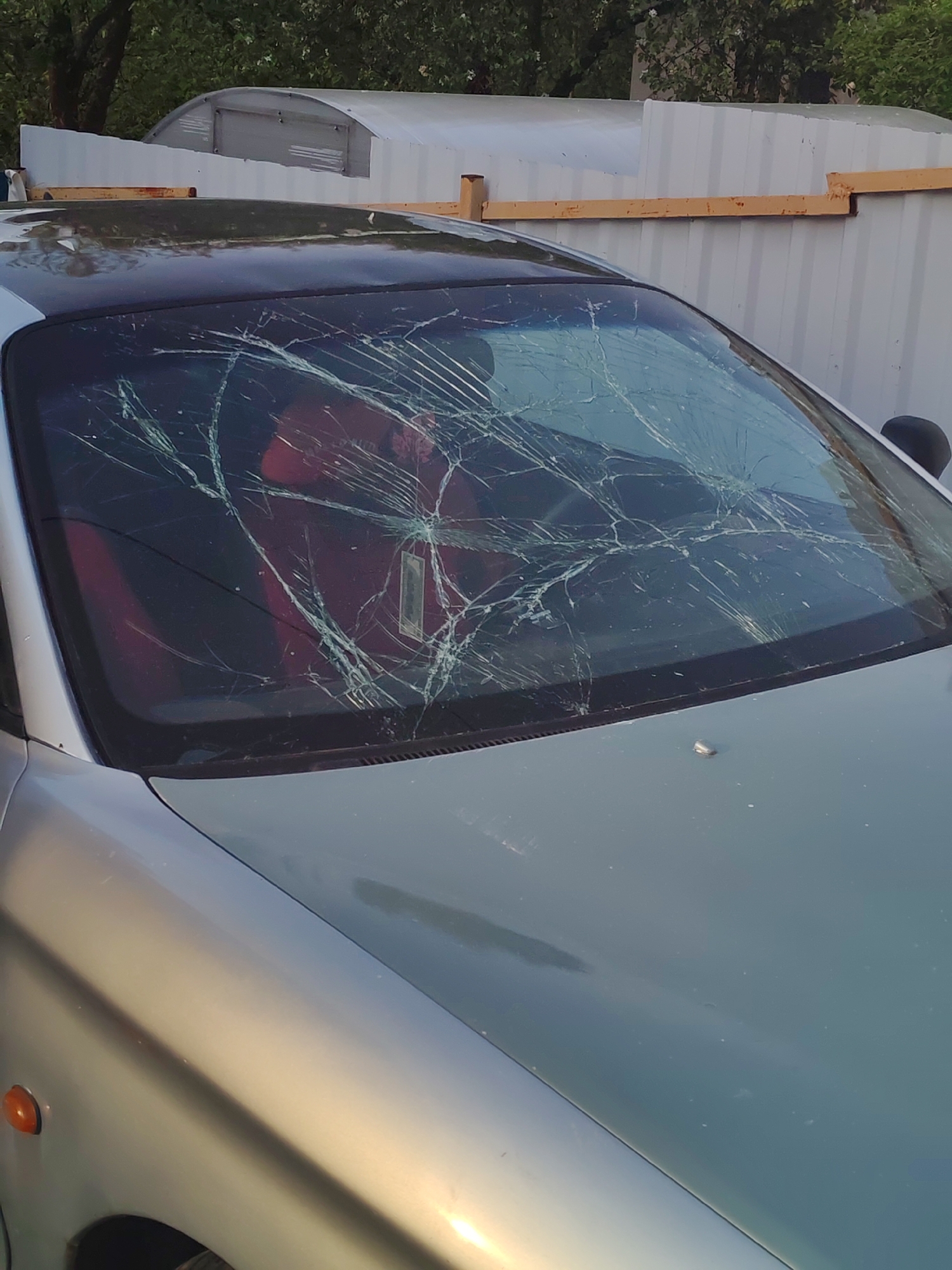 The image depicts a silver car with a severely cracked windshield. The interior has red seats. In the background, there's a corrugated metal structure with wooden supports. The setting appears to be an outdoor location in a city.