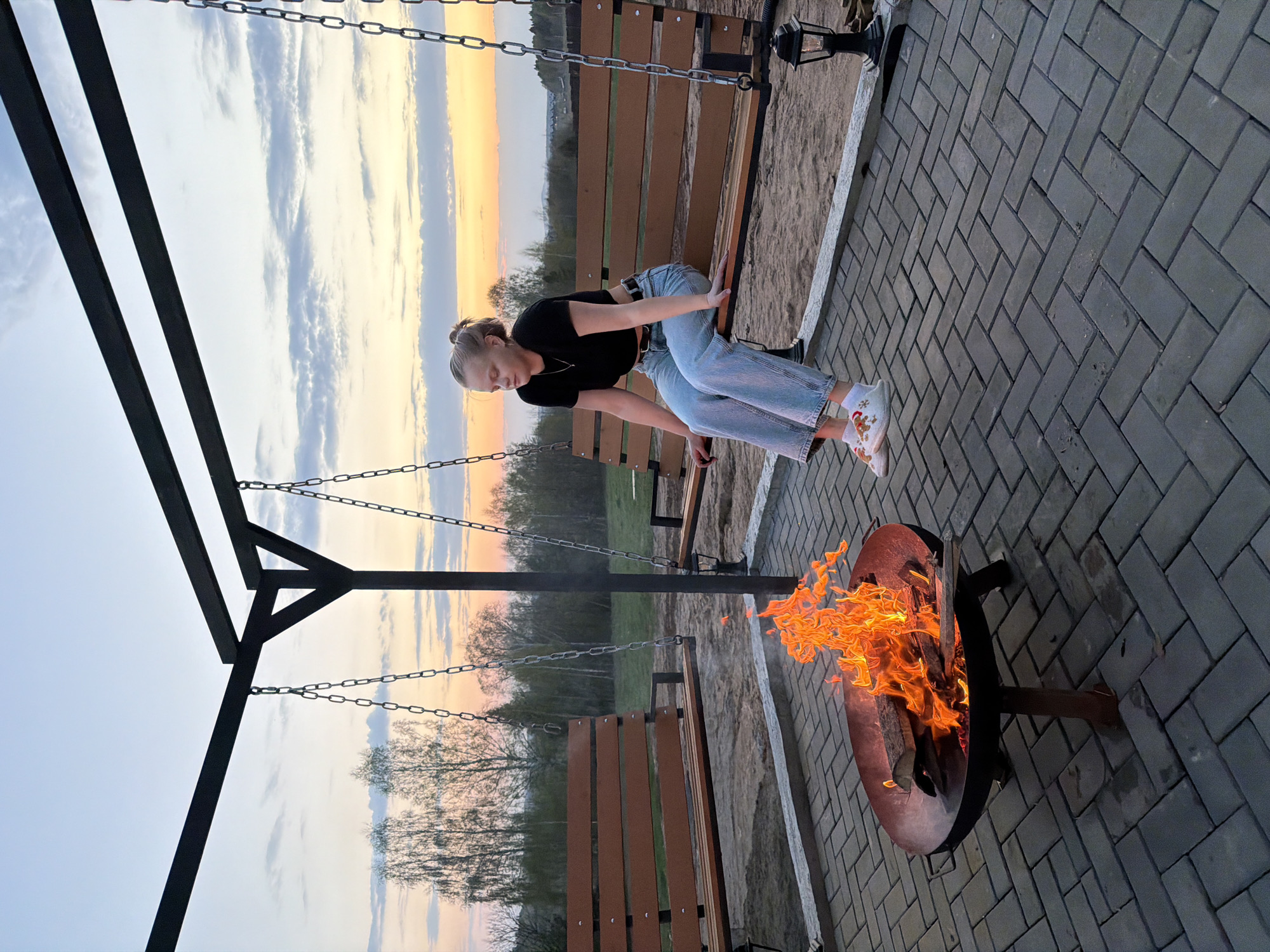 The scene shows a young woman sitting on a swing next to a fire pit. The woman has blonde hair, wears a black shirt, light blue jeans, and white shoes. She is leaning forward with her hands on the swing seat. The fire pit contains a large fire with bright orange flames. In the background, there is a fence, trees, and a sky with colors ranging from blue to orange, indicating either a sunrise or sunset. The ground is paved with gray bricks in a herringbone pattern. The location may be in Kamennoye, Russia.