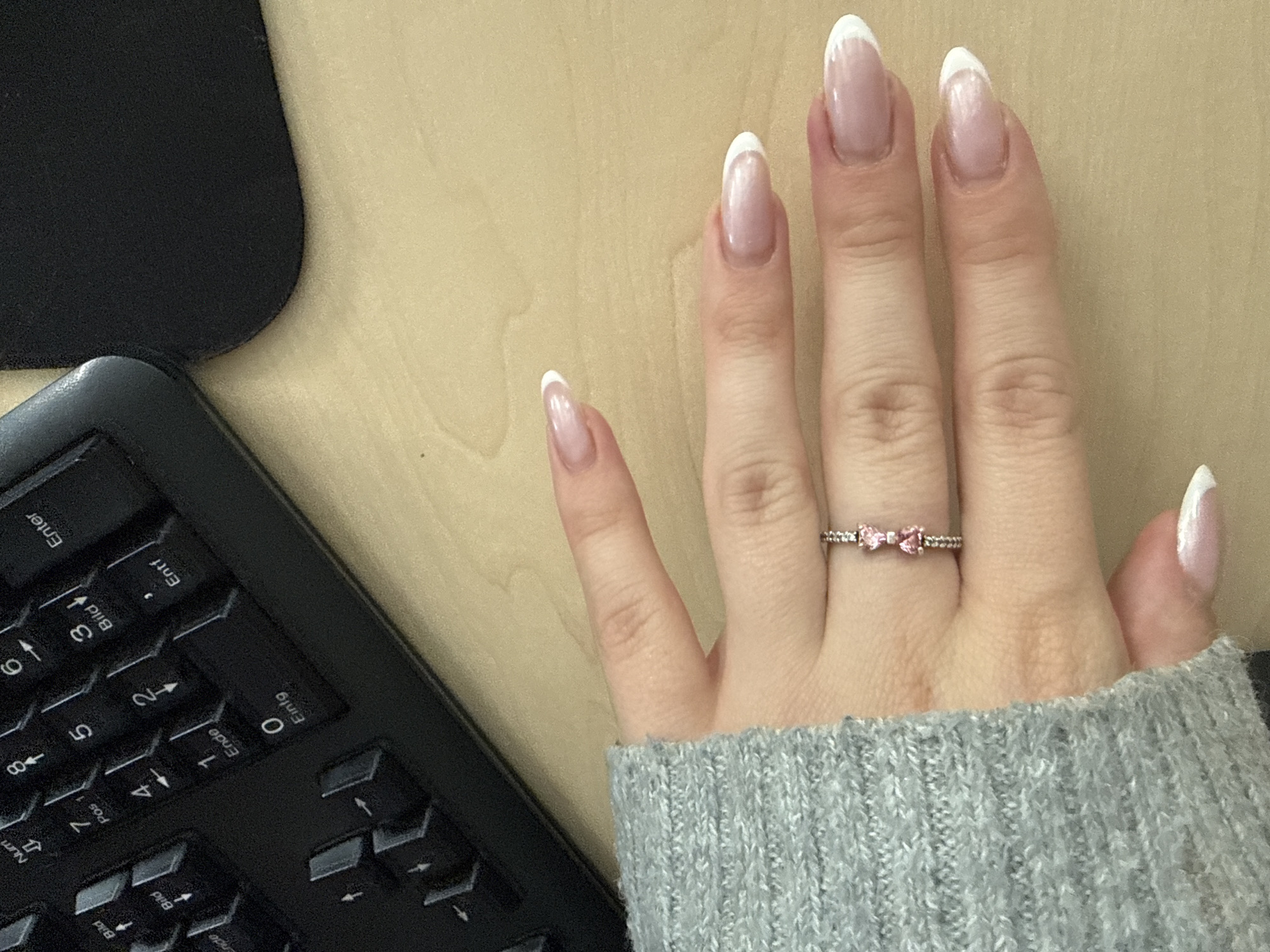 In a close-up, overhead shot on a light wood desk, a woman's left hand is positioned with her fingernails facing upwards. She is wearing a grey knitted sweater on her arm. On her ring finger, a silver ring with two small pink heart-shaped stones is visible. The woman's fingernails are manicured with a French tip style, featuring a light pink base and white tips. To the left of her hand, a portion of a black keyboard is visible, with keys labeled in German, including "Bild," "Pos," "Ende," "Einfg," and "Enter." A black object, possibly part of a laptop or other electronic device, is seen in the upper right corner of the frame. The overall lighting suggests an indoor, possibly office environment.