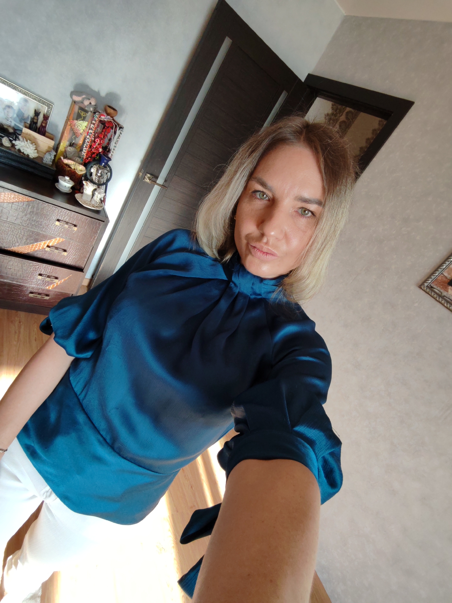 A woman with blonde hair and green eyes is captured in a selfie taken indoors, likely in Petropavlovsk-Kamchatsky, Russia. She wears a teal blue, high-necked blouse with voluminous sleeves and white pants. Her expression is neutral, with a slight pout, as she looks directly into the camera held at a low, upward angle.
The background reveals an interior space, possibly a bedroom or living room. To the left, a dark brown dresser with a crocodile skin texture on its drawers and metallic handles is visible. On top of the dresser, various decorative items are arranged: a framed picture containing multiple figures, small figurines (including what appears to be a black cat), a wooden stand displaying an assortment of red and black beaded necklaces or bracelets, small white teacups or bowls, and other trinkets.
Behind the dresser, a dark brown door with a metallic handle is partially open. Another dark framed opening, possibly another door or a mirror, reflects a pattern on the wall or a window. The walls are a light, possibly textured, neutral color. The floor is made of wood, and sunlight casts strong highlights and shadows, suggesting it is daytime. No visible text is present in the image.