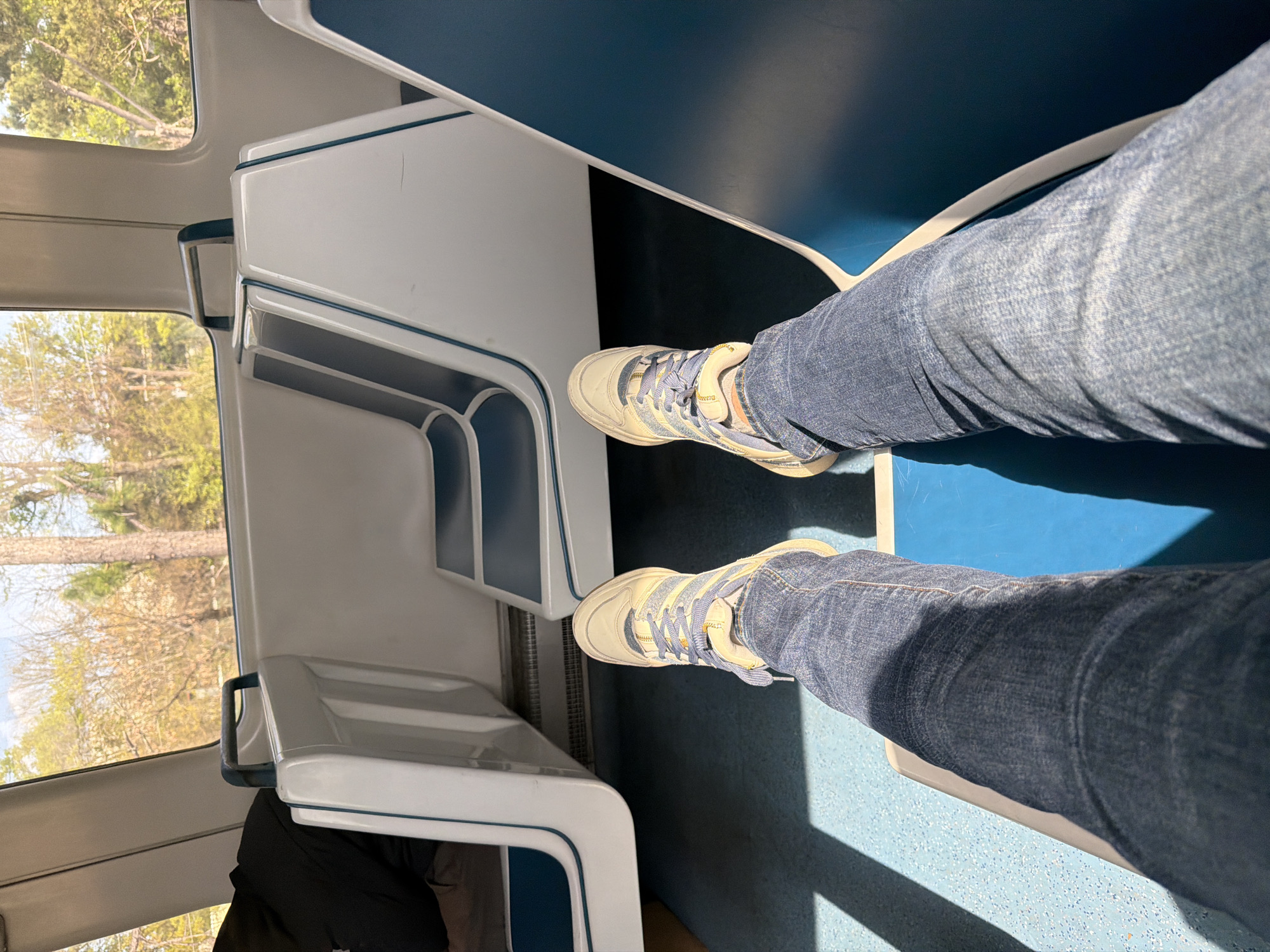 The image shows a person seated on public transportation, specifically the MARTA (Metropolitan Atlanta Rapid Transit Authority) train in Atlanta, Georgia, United States of America. The individual is wearing blue jeans and tan sneakers with blue laces. The interior of the train car features white and blue seating. Trees and sky are visible through the window. A dark object sits in the bottom left, suggesting a bag or clothing. The flooring is a blue speckled pattern. Strong shadows from overhead light are visible. The angle is a first-person perspective.