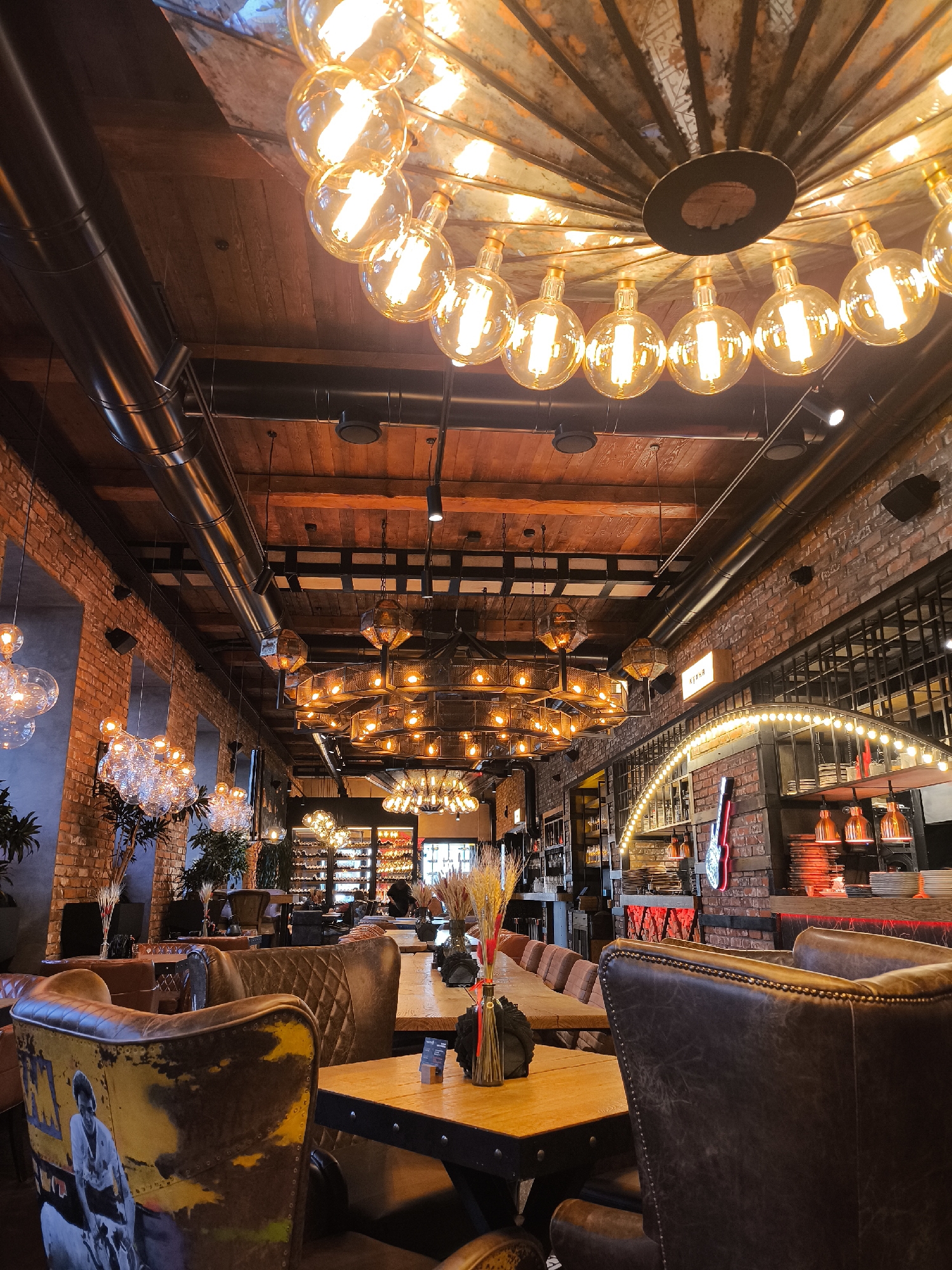 This image depicts the interior of the "Кино" (Kino) restaurant, located at ulitsa Kirova, 107, Izhevsk, Russia. The scene presents a high-ceilinged, industrial-style dining or bar area. Reddish-brown exposed brick walls with tall, dark-framed window cutouts line the left side, while the ceiling features dark wooden planks with visible metallic ventilation ducts and track lighting.
Numerous lighting fixtures illuminate the space. Prominently, a large, ornate chandelier with multiple spherical filament bulbs hangs centrally in the foreground. Further down the long room, a series of similar, but smaller, multi-bulb chandeliers are suspended. On the left, clusters of clear glass sphere lights are visible. On the right, geometric metal cage chandeliers and an illuminated archway with string lights accent a bar area.
The furniture consists of dark brown upholstered seating, including armchairs, some with distressed leather finishes and one in the foreground displaying a black and white graphic print of a person. Wooden tables are arranged in rows, some set with dark vases containing dried wheat stalks. The bar area on the right features stacked white plates, copper-toned pendant lamps, and a red neon guitar sign. In the background, a large display case with shelves, possibly for bottles, is visible. Potted plants are positioned throughout the interior. No individuals are clearly visible or engaged in specific actions within the frame.