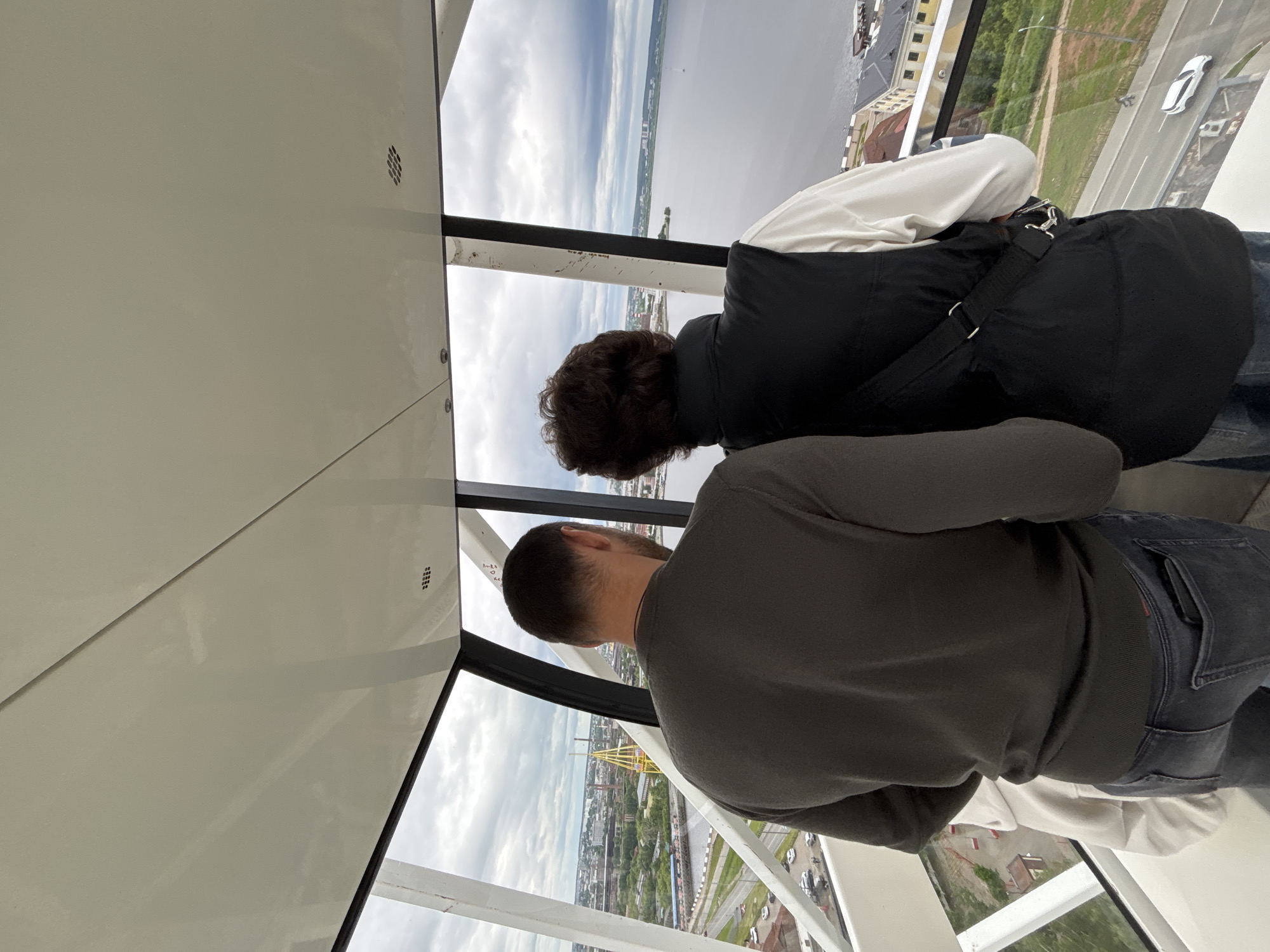 The image captures an interior view of an observation cabin, likely part of a Ferris wheel, with two individuals seen from behind looking out large windows at the city of Izhevsk, Russia. The cabin's interior features a light off-white ceiling and structural elements, including visible seams and small rectangular grilles on the ceiling. The windows are framed by white and dark vertical and diagonal supports, revealing an expansive view.
The person on the left, a male, wears a dark olive green long-sleeved top and dark jeans, with a phone partially visible in his right back pocket. A light-colored garment is partially visible behind his left arm. The person on the right, with dark, wavy hair, wears a white long-sleeved shirt under a black puffer vest and carries a black cross-body bag with a silver buckle. Both individuals are standing, gazing out at the urban and natural landscape.
Through the left window, a sprawling city is visible, characterized by various buildings, industrial structures, and green areas. A distinctive yellow, lattice-like Ferris wheel, identified as the "Zolotoy Ostrov" (Golden Island) Ferris wheel, is a prominent landmark in the middle distance. Roads with parked cars and a narrow body of water with small boats are also visible. Through the right window, a vast expanse of calm, grey water, confirmed as the Izhevsk Pond, stretches to the horizon where distant land and buildings are discernible. Immediately below, older buildings with yellow facades and dark roofs line a road with a white car and a pedestrian. The sky above is heavily overcast with grey clouds.