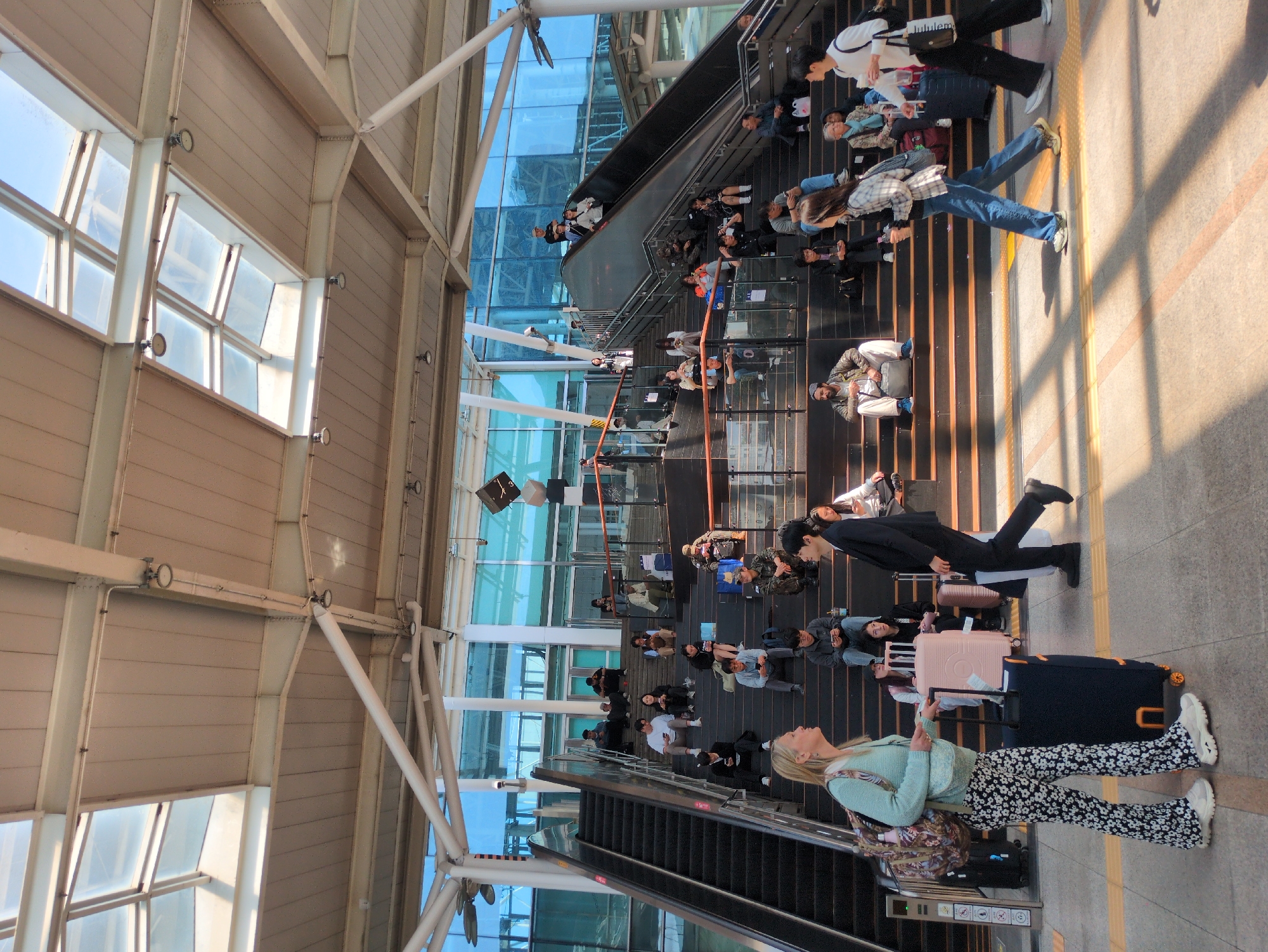 The image shows the interior of what appears to be a train station or transportation hub in Seoul, South Korea. The architectural style features high ceilings with metal beams and large glass windows, allowing natural light to flood the space. A large glass wall is a prominent feature, running vertically up the entire space. Numerous people are visible, many of them seated on the ground or stairs, appearing to be waiting. Luggage, including wheeled suitcases, is seen scattered among the people, indicating they are likely travelers. An escalator is also visible. The general atmosphere is one of a busy, transit environment filled with people.