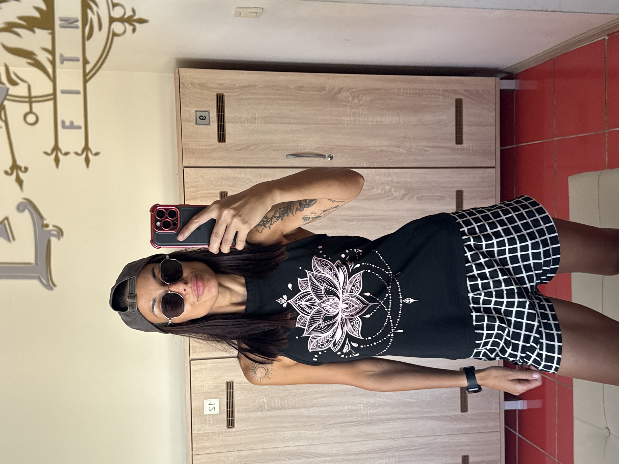 A woman wearing a black tank top with a lotus flower design, black and white checkered shorts, sunglasses, and a grey baseball cap is taking a mirror selfie in a locker room. She is holding a smartphone in her right hand. The locker room has wooden lockers and a red tiled floor. Above the lockers, a decorative "FITNESS" sign is visible. The image appears to be taken in Pleven, Bulgaria, based on the context provided in the prompt, though no explicit visual indicators within the image itself confirm this location.