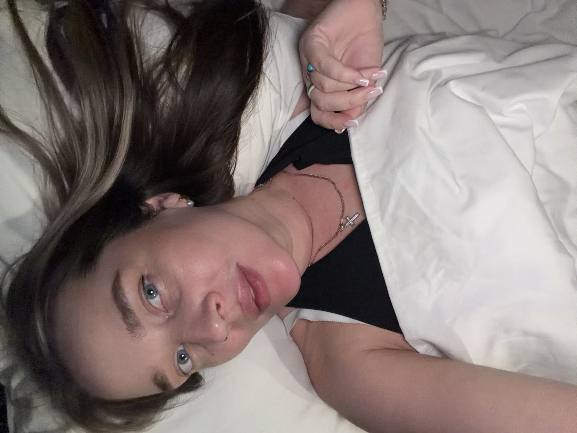 A medium close-up image captures a person lying down on white bedding, partially covered by a white sheet or blanket. The subject is a female with long, dark brown hair featuring lighter brown or blonde highlights, which is spread out on the pillow to the left. She is positioned with her head tilted, looking directly towards the camera.
Her facial features include blue eyes, prominent eyebrows, and a small silver hoop piercing in her left nostril. Her lips are slightly parted, revealing a neutral to slightly pensive expression. She wears a black top or camisole visible beneath the white bedding. A thin, rose-gold or gold-toned chain necklace with a small, ornate cross pendant rests on her collarbone.
Her left hand is visible, resting near her head, showing a French tip manicure on her nails. She wears several rings: a noticeable turquoise stone ring on her ring finger, and other silver-toned rings on her middle and possibly index fingers. A small, silver-toned earring is visible in her left ear.
The scene appears to be an indoor setting, likely a bedroom, suggesting a moment of rest or relaxation. No specific time of day or weather conditions can be discerned from the image. While the provided context indicates Moscow, Russia, there are no visual cues within the image itself to confirm this location. No text is visible in the media file.