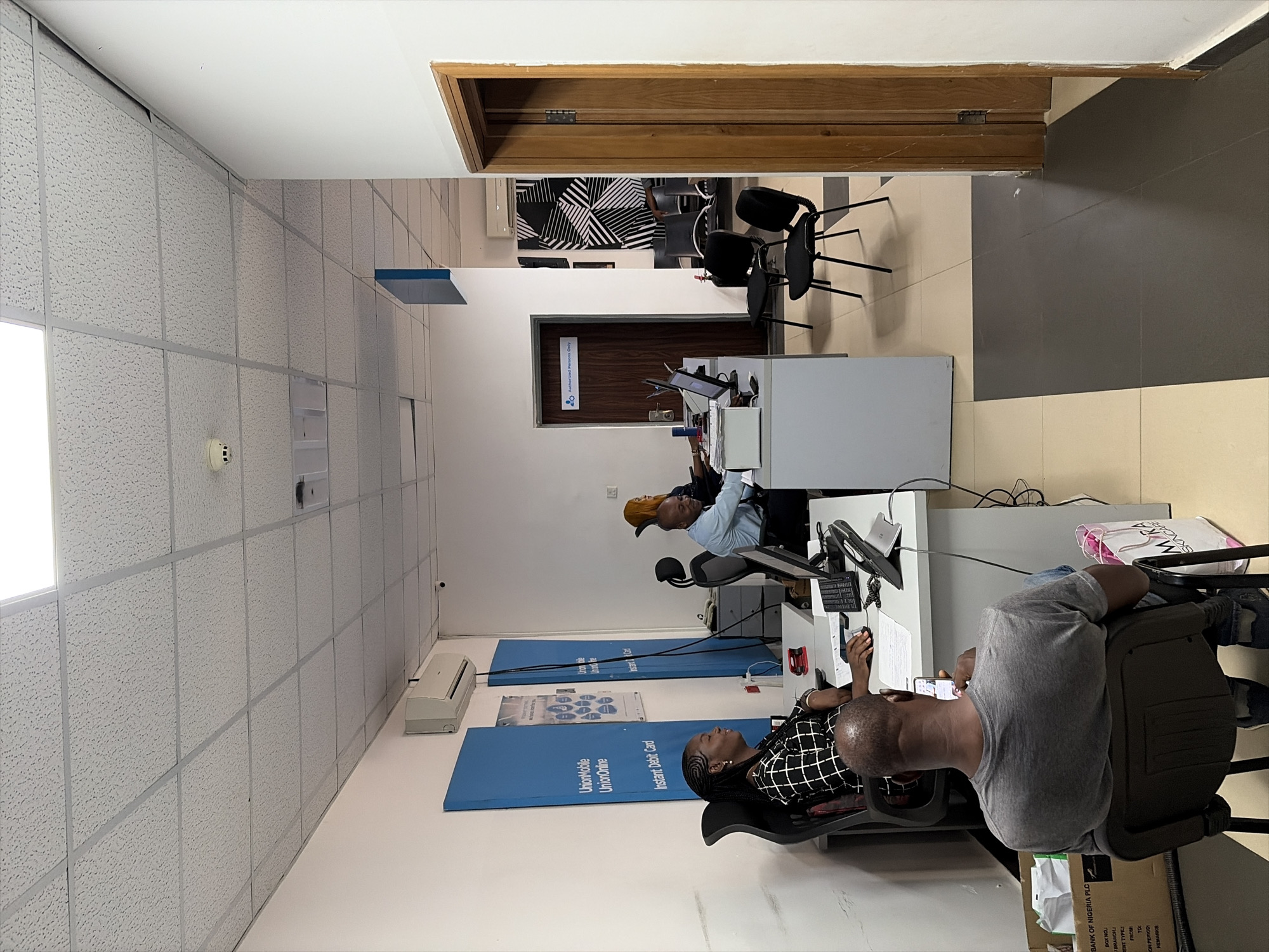 This image captures an indoor scene, likely a bank branch or customer service office, located in Abuja, Nigeria. Several people are present, appearing to be engaged in work or waiting. In the foreground, a man in a grey t-shirt sits with his back to the camera, looking at his mobile phone. Beside him, a woman in a black and white checkered top sits at a white desk, facing forward. Further back, another man in a light blue shirt is seated at a desk, focused on a computer screen.
The environment is furnished with multiple white office desks, each equipped with computer monitors and keyboards. Office chairs are arranged for both staff and possibly customers. An air conditioning unit is visible on a wall. Prominently displayed are two blue banners promoting "Union Mobile," "UnionOnline," and "Instant Debit Card," reinforcing the banking context.
Notable details include a dark wooden door in the background with a sign that reads "Authorized Persons Only." A cardboard box on the floor near the man in grey displays partial text including "BANK OF NIGERIA PLC," "BRANCH:", "BENT TYPES," "FROM," "TO:", "ON PERIOD," and "REMARKS." A white bag with "SKINCARE MIRA" printed on it is also visible. The walls are light-colored, featuring sections with acoustic tiles. The ceiling consists of standard dropped tiles, and a fire alarm is mounted on a wall. The background shows a section with a black and white abstract pattern, likely wallpaper or a partition. The space is brightly lit by overhead lighting, suggesting daytime.