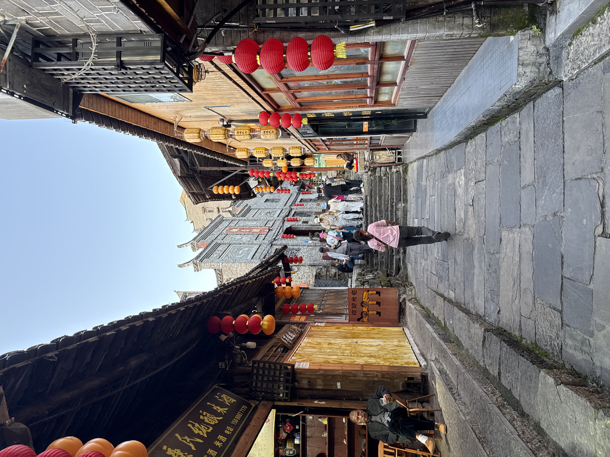 The image depicts a narrow, stone-paved street in Furong, China. Red and yellow lanterns are strung above, lining the street. Traditional Chinese buildings flank the street, with intricate wooden structures. People are seen walking up stairs in the distance. An older man is seated in front of a shop on the right, while other people are walking the street. The sky is pale blue, partially visible at the top.