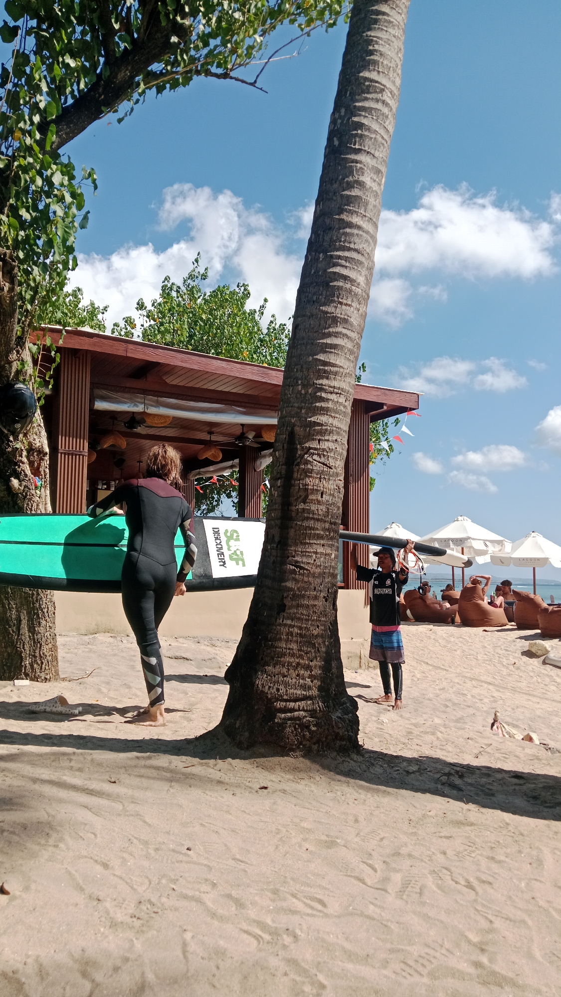 In Kuta, Indonesia, on a sandy beach under a clear blue sky with scattered clouds, a person wearing a black and maroon wetsuit walks towards the left carrying a teal surfboard. The surfboard has "Surf Discovery" written on it. To the right of the wetsuit-clad individual and partially obscuring the view, a large palm tree trunk stands tall. Further to the right, another person, wearing a black t-shirt and striped shorts over leggings, holds a surfboard overhead. In the background, a wooden building with a red roof functions as a bar or restaurant, with fans visible and several people lounging on beanbag chairs under white umbrellas. The environment suggests a recreational setting, likely a surf school or beach resort.